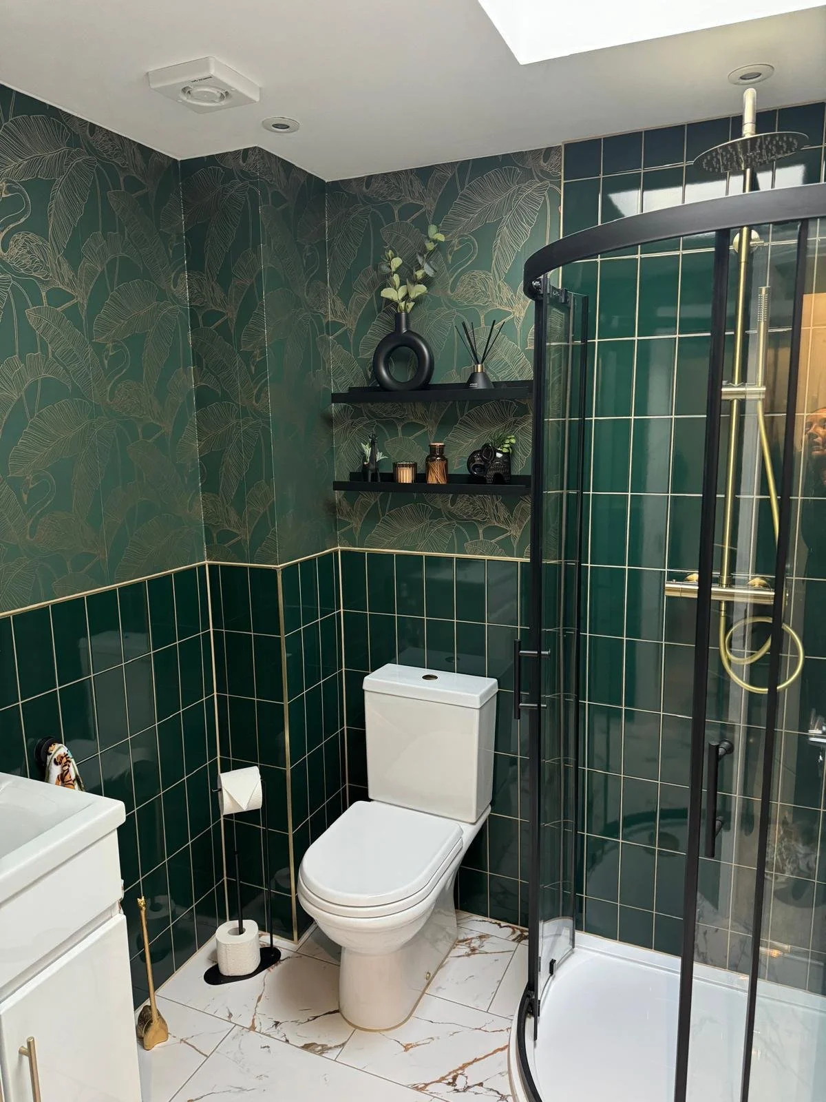 Modern bathroom with green leaf-patterned wallpaper, black shelves with decorative items, white toilet, shower enclosure with gold fixtures, and white marble floor tiles.