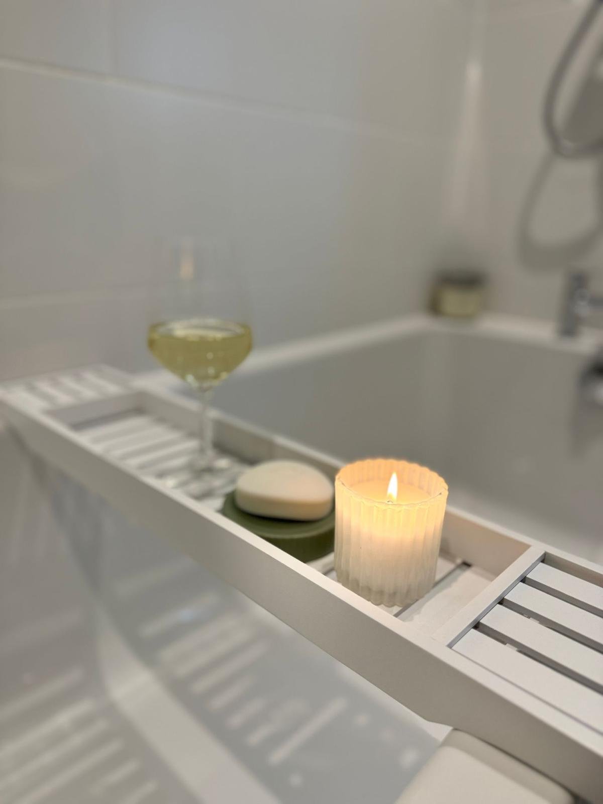 Close-up of a white bathtub with a bath tray holding a lit candle, a bar of soap, and a glass of white wine, in a bathroom.