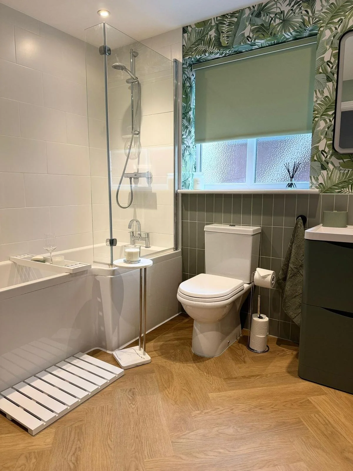A modern bathroom with a walk-in shower, bathtub, toilet, and storage cabinet. The bathroom features green foliage-patterned wallpaper and a window with a green roller shade. The floor has wood-look vinyl flooring.