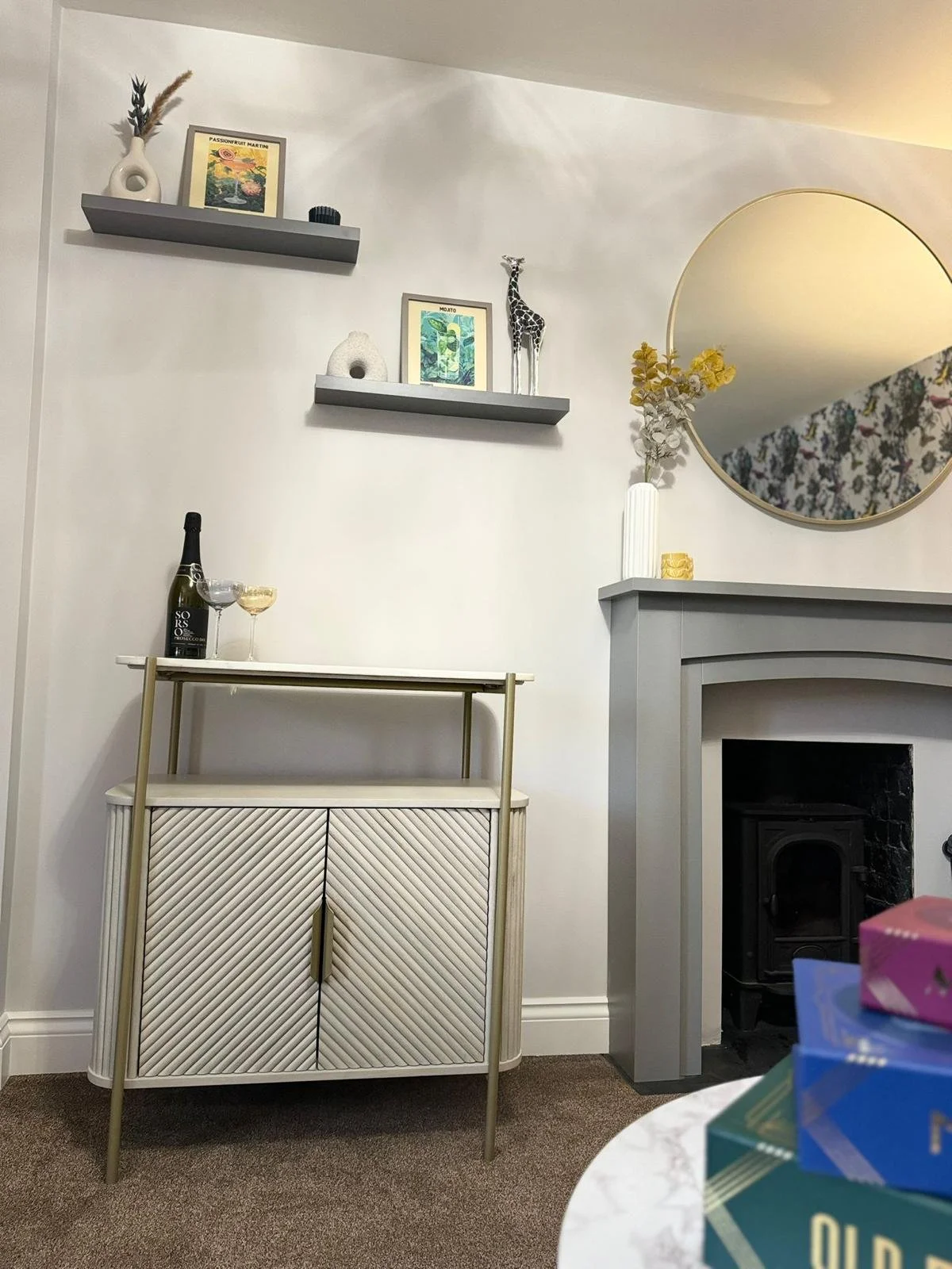 Decorative living room corner with a white cabinet, a small gold bar cart with a bottle of wine and wine glasses, two gray floating shelves with art and decorative items, a round mirror on the wall, a fireplace mantel with a vase and flowers, and col