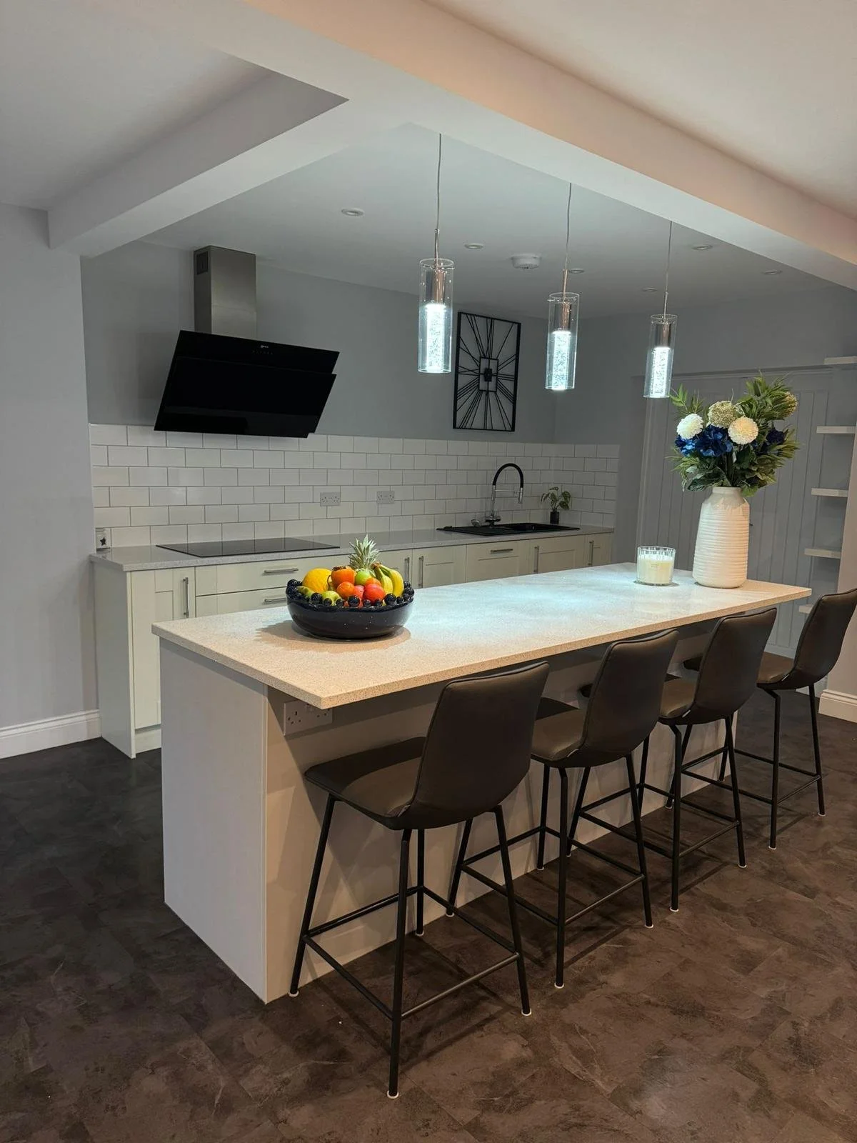 Modern kitchen with a white countertop island, black bar stools, pendant lights, a bowl of fruit, a vase of flowers, and a wall clock.