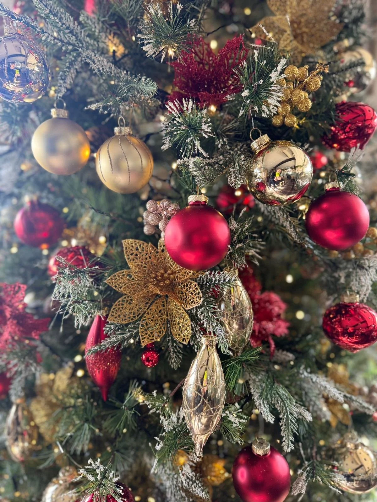 Close-up of a decorated Christmas tree with red, gold, and silver ornaments, gold poinsettia and star decorations, and snow-like flocking on the branches.