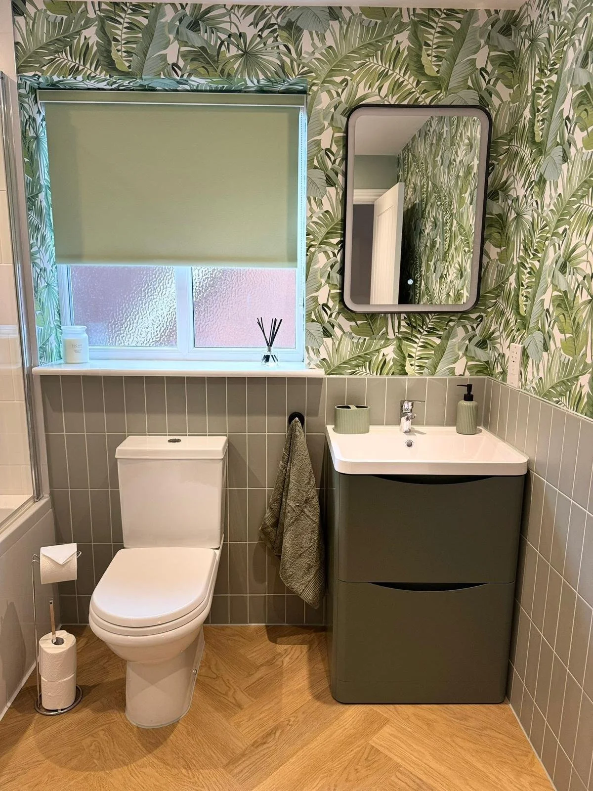 A bathroom decorated with green tropical leaf wallpaper, a frosted window with a green roller shade, a mirror, a white toilet with a toilet paper roll on a stand, a small gray towel hanging, a green and white vanity sink with a faucet, a soap dispens