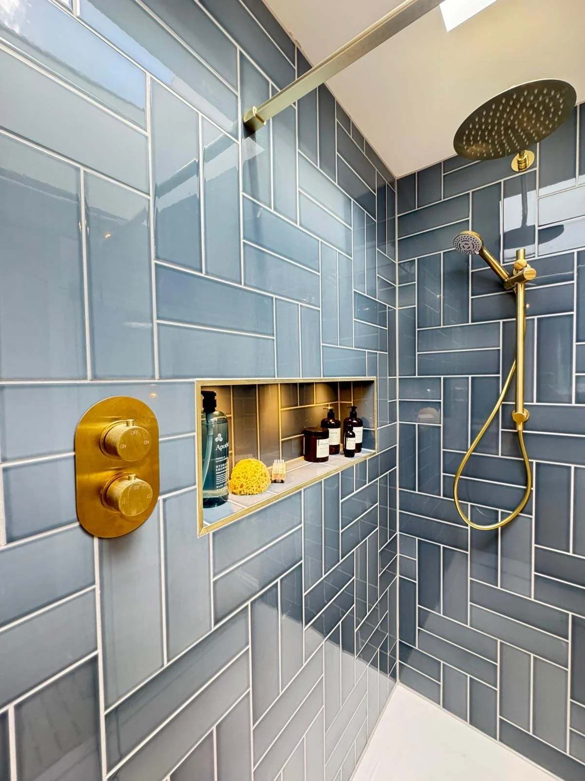Modern shower with blue subway tiles, gold shower hardware, a built-in niche with toiletries and a yellow sponge, and a ceiling-mounted rainfall shower head.