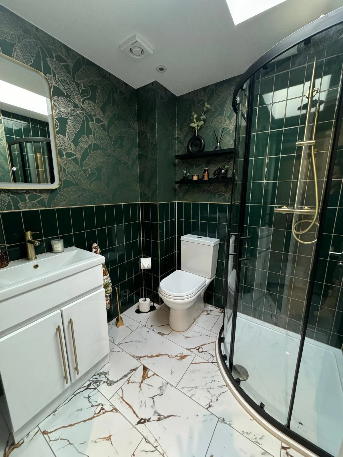 Modern bathroom with dark green tiled walls, white marble floor, and a glass shower enclosure. Features include a white vanity with gold handles, a small mirror, a toilet, and wall-mounted shelves with decorative items.
