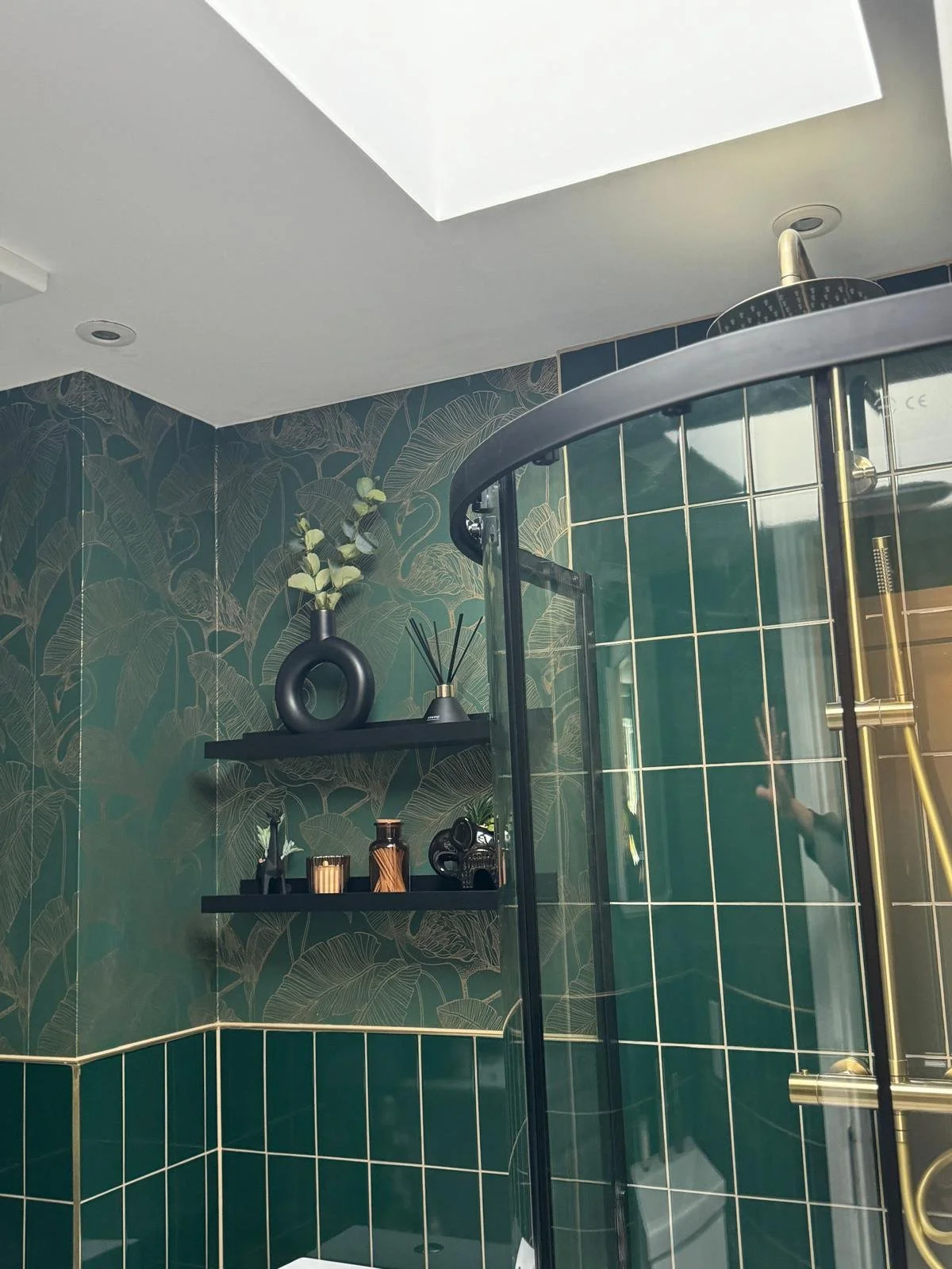 A modern bathroom shower area with dark green tiles and tropical leaf patterned wallpaper, featuring black shelves with decorative objects and a brass shower fixture.