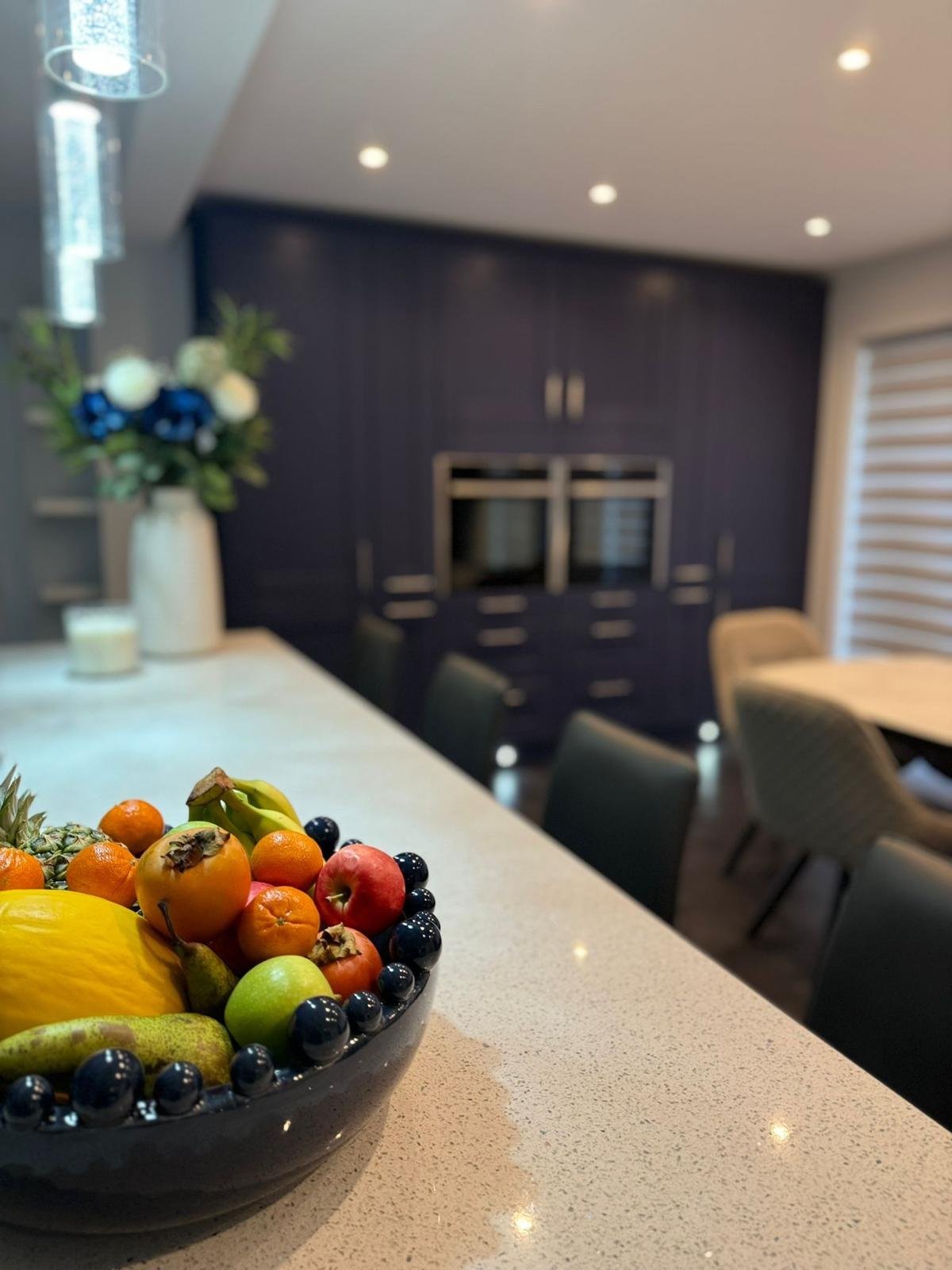 A black bowl filled with assorted fruits, including apples, oranges, bananas, pineapple, and grapes, on a white countertop in a modern kitchen or dining area.