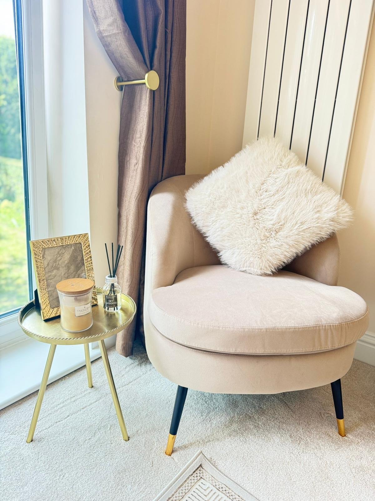 A cozy indoor corner with a beige velvet armchair with a fluffy white pillow, next to a small round gold side table holding a candle, picture frame, and diffuser, near a window with purple curtains.
