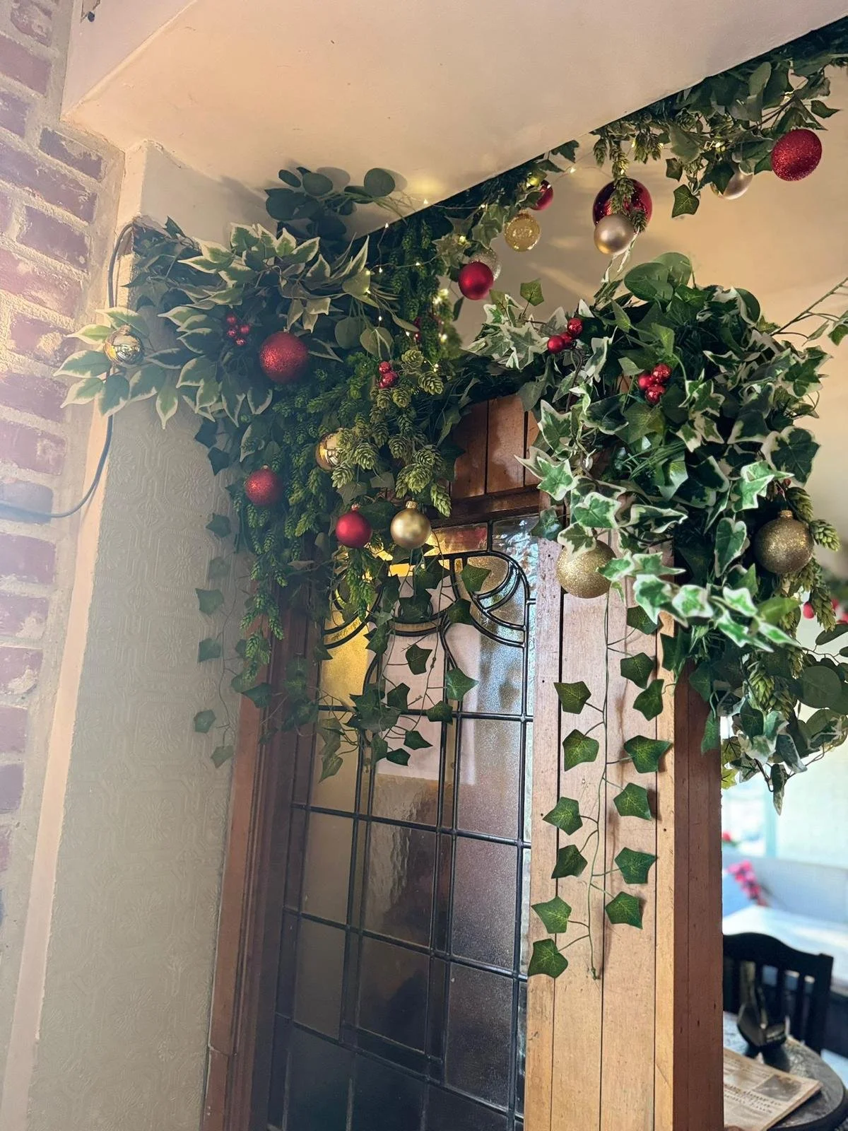 Decorative holiday greenery with red and gold ornaments hanging above a window with a wooden frame and iron grid, inside a cozy room.
