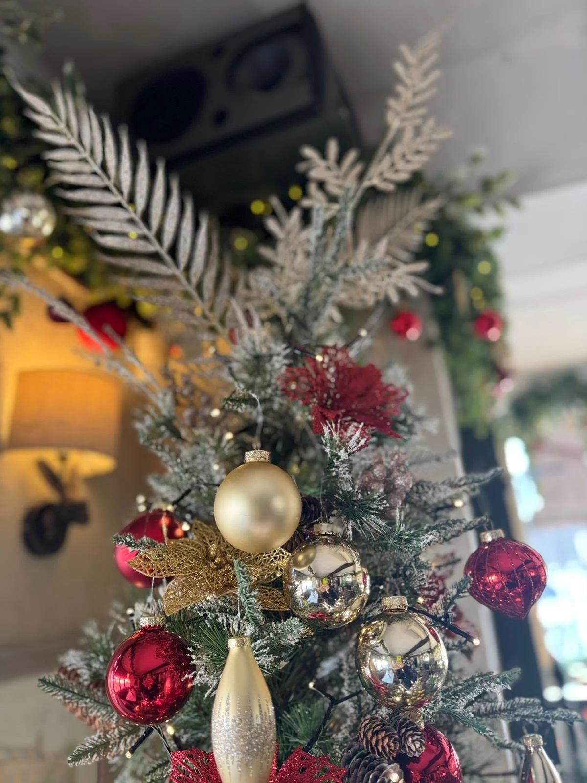 Close-up of a decorated Christmas tree with red, gold, and silver ornaments, pinecones, and a gold glittery leaf, with a blurred background of a room with warm lighting.