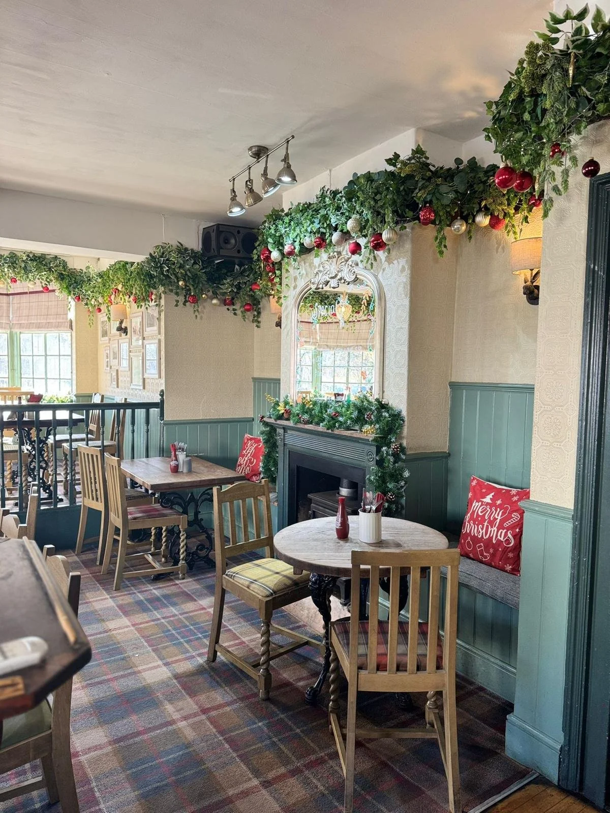 A cozy restaurant decorated for Christmas with garlands, baubles, and wreaths. Wooden tables and chairs are arranged on a plaid carpet, with holiday cushions on the benches. The room has natural light coming through windows and a fireplace with holid