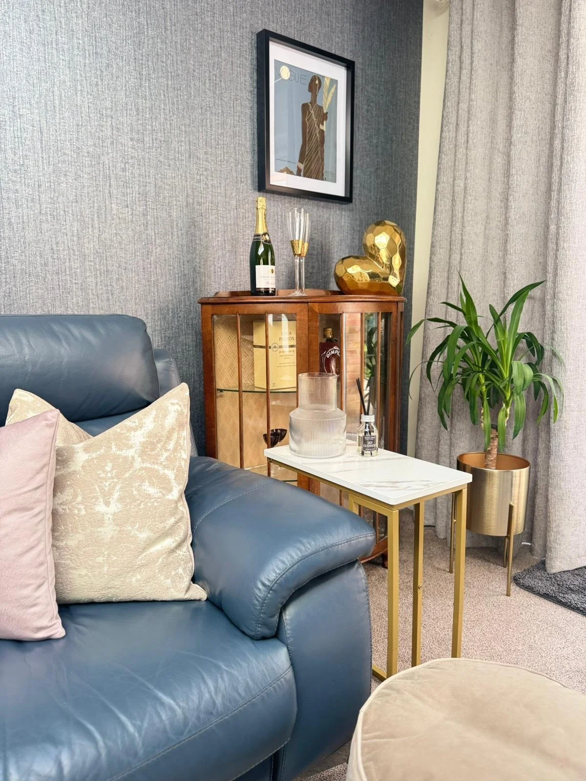 A living room corner with a blue leather sofa, beige pillows, a white and gold side table, a wooden cabinet with glass doors, a potted plant, and framed artwork on the wall.