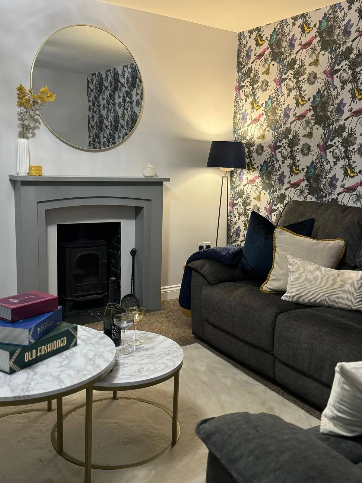 A cozy living room with a gray sofa and several decorative pillows, a white marble coffee table with books and wine glasses, a fireplace with a round mirror above, a black floor lamp, and floral wallpaper with birds on the wall.