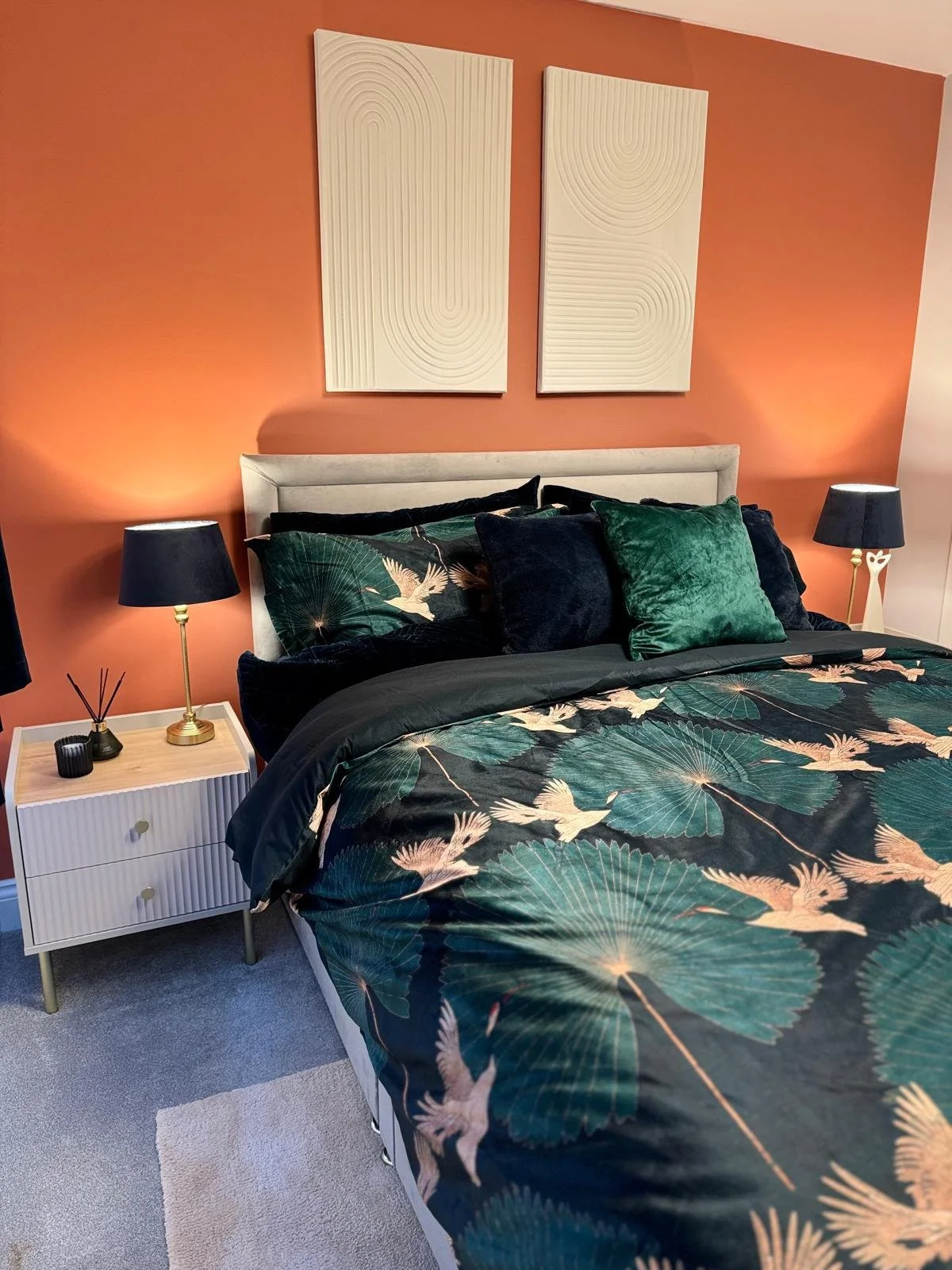 Bedroom with an orange accent wall, two white abstract wall art pieces, a bed with dark green, black, and patterned bedding featuring cranes and leaves, black and green pillows, white nightstands with black lamps and decor, and light gray carpet.