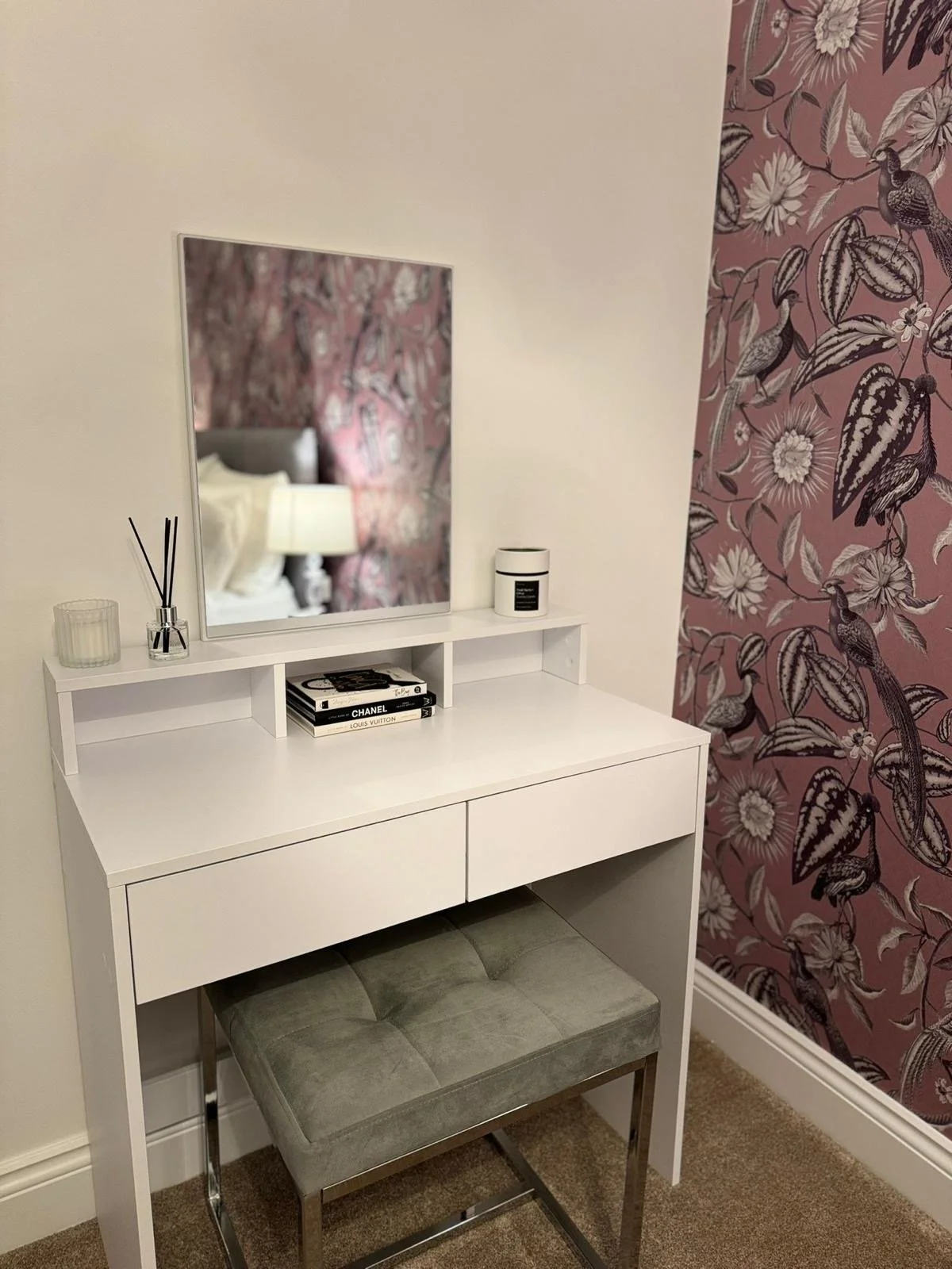 A white vanity table with a mirror, a candle, a glass container, a small tray with books, and a vanity light. A cushioned stool with metal legs is placed in front. The wall behind has pink floral and bird wallpaper.