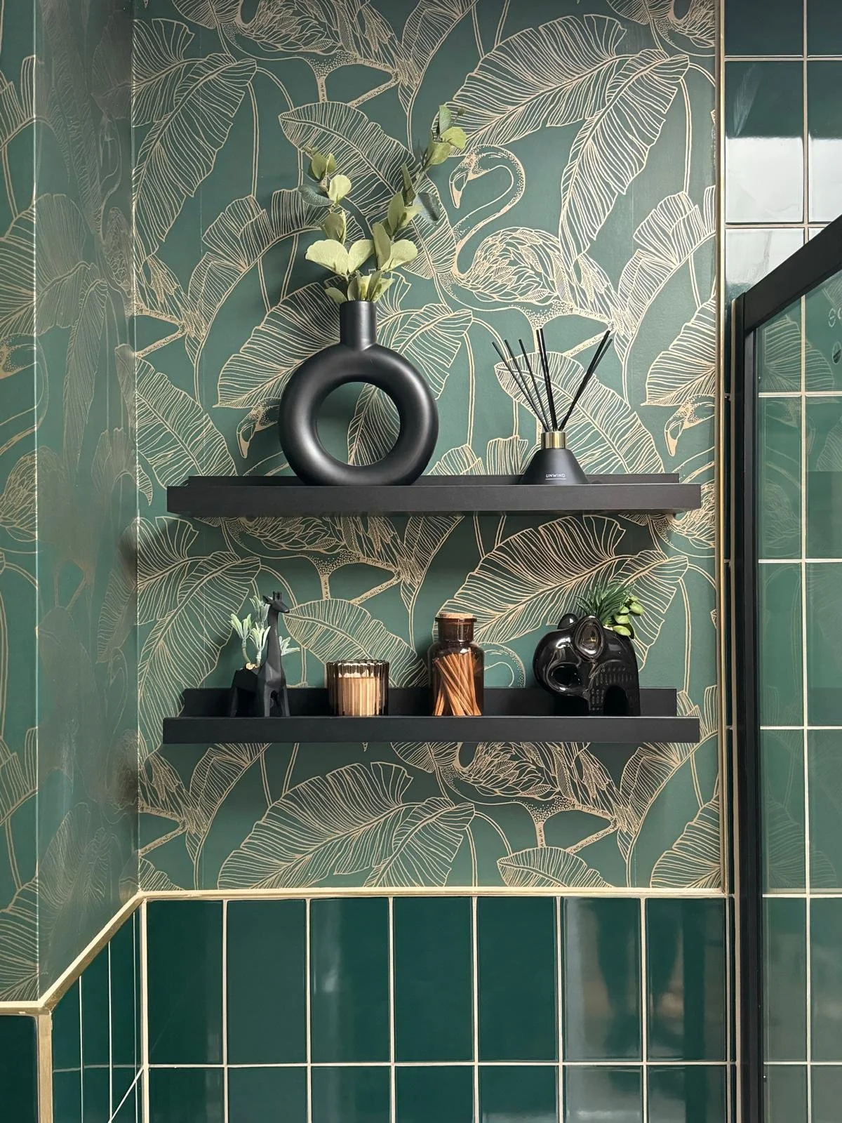 Decorative bathroom corner with green leaf patterned wallpaper, dark green tiled walls, and two black floating shelves holding black and gold vases, a small decorative giraffe, a black elephant head sculpture, and green plants.