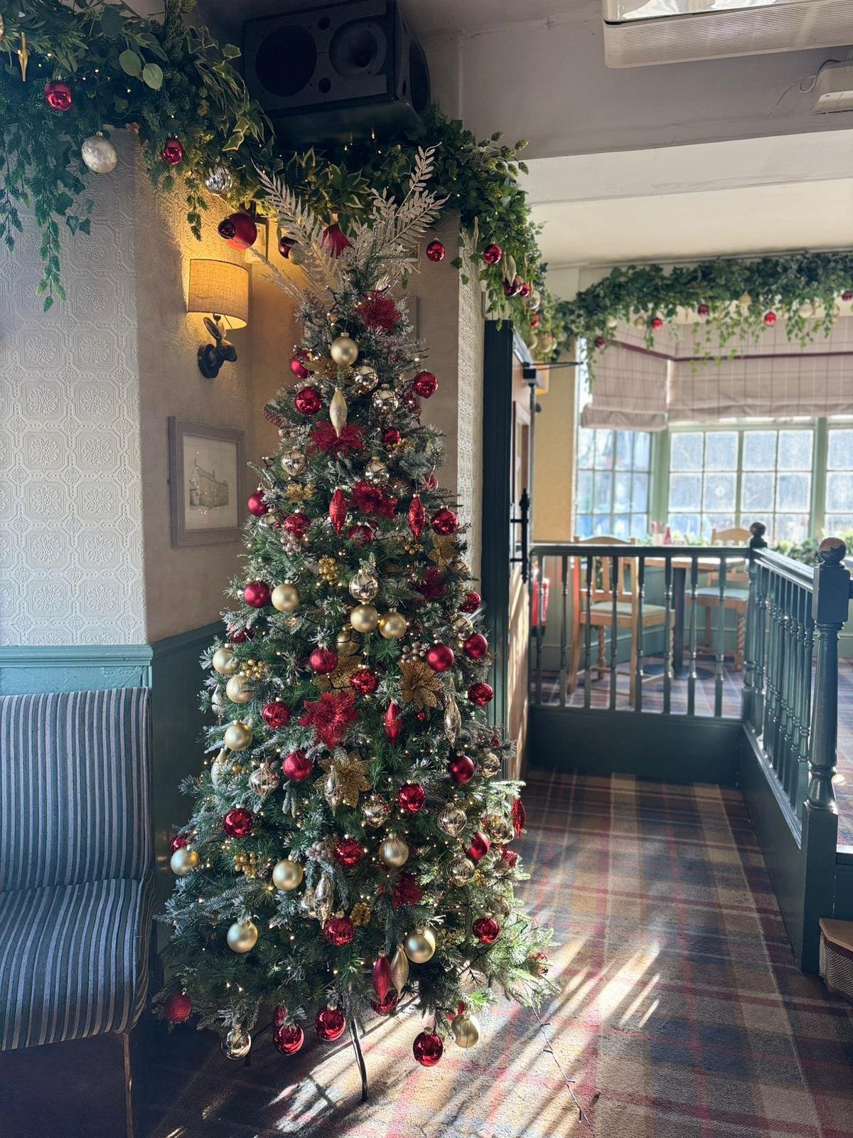 Decorated Christmas tree with red, gold, and silver ornaments in a cozy, well-lit restaurant or cafe with holiday garlands and a window in the background.