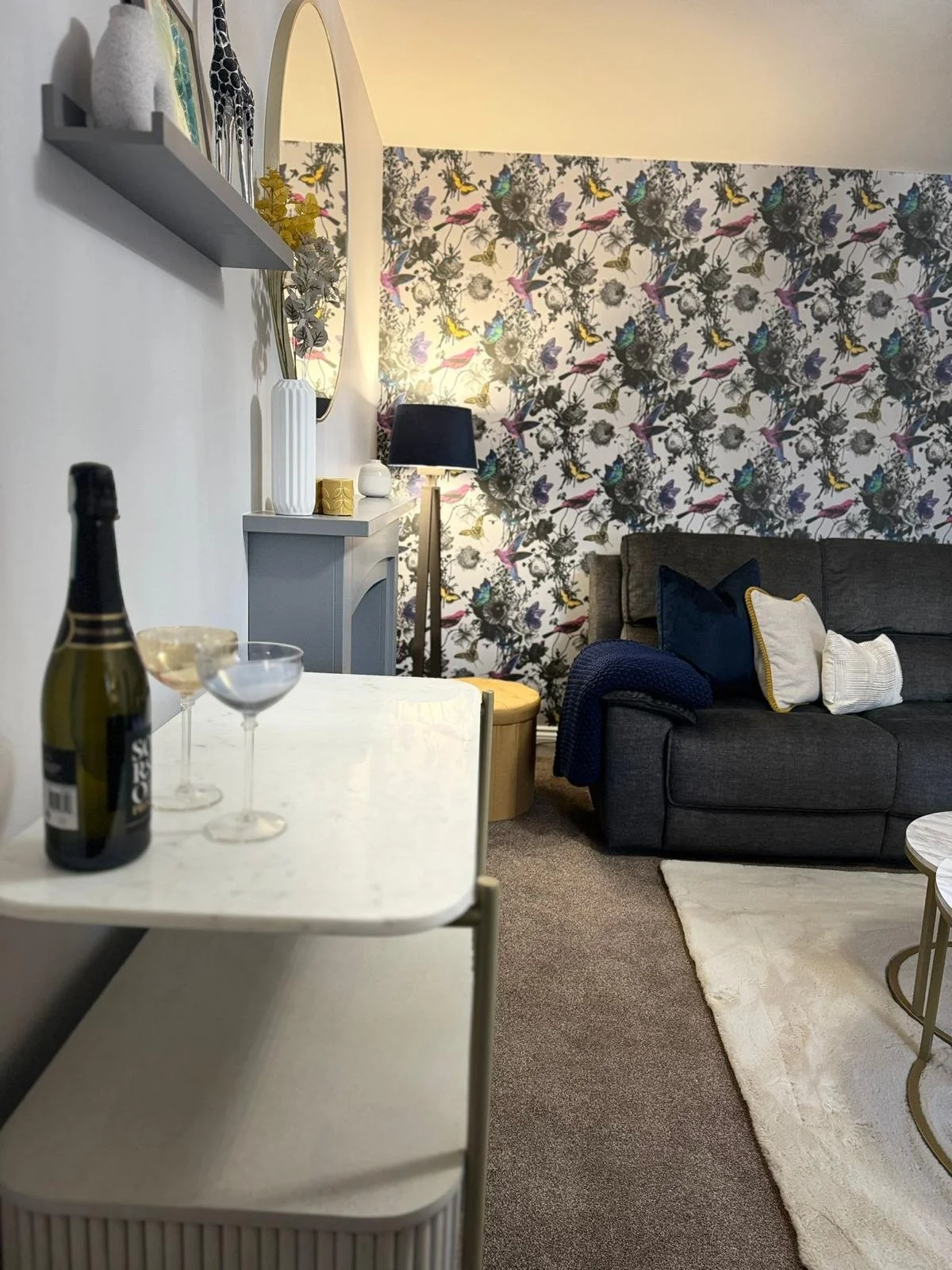 A living room with a dark gray couch, a patterned floral wallpapered wall, a white shelf with vases and decorative items, a floor lamp with a black shade, and a small table with a marble top and gold frame. There is a small curtain-free window next t