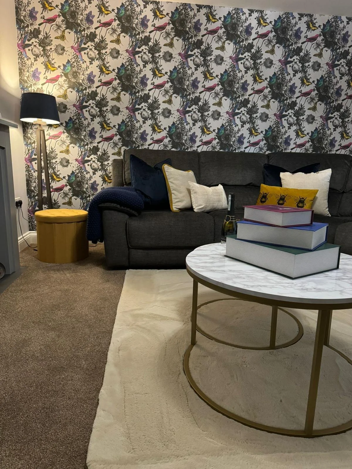 Living room with floral and bird patterned wallpaper, dark sofa with pillows, a white rug, a round marble coffee table with stacked books, a yellow ottoman, and a black floor lamp.