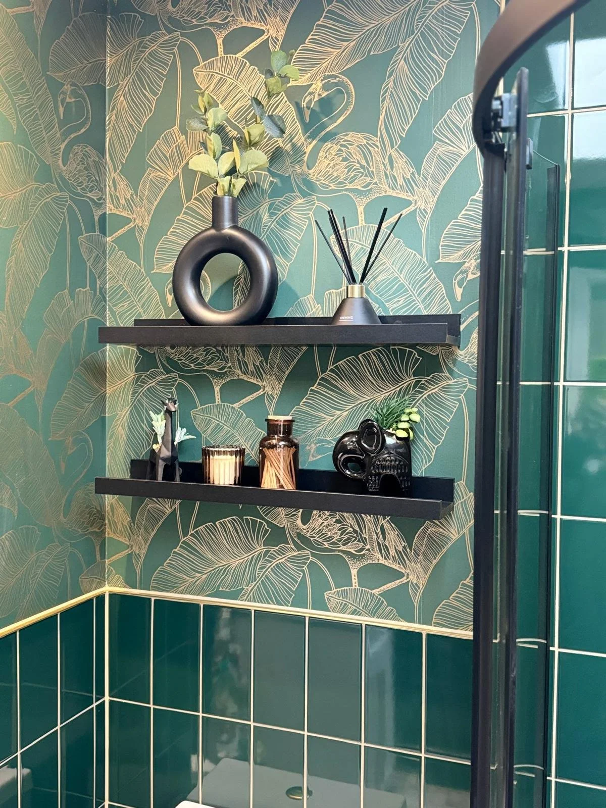 Decorative bathroom wall with tropical leaves wallpaper, black floating shelves holding a black vase with greenery, reed diffuser, black skull ornament, small plant, and figurines, with green tiled lower wall.