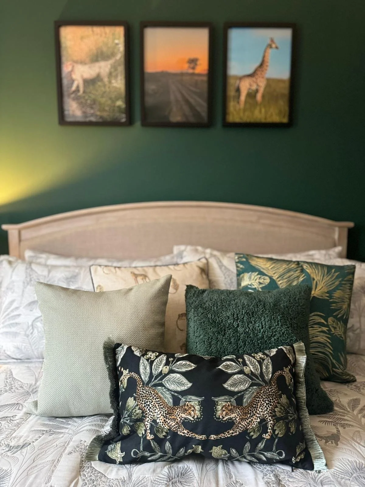 A bed with multiple decorative pillows in front of a green wall and three framed landscape photographs above the headboard.