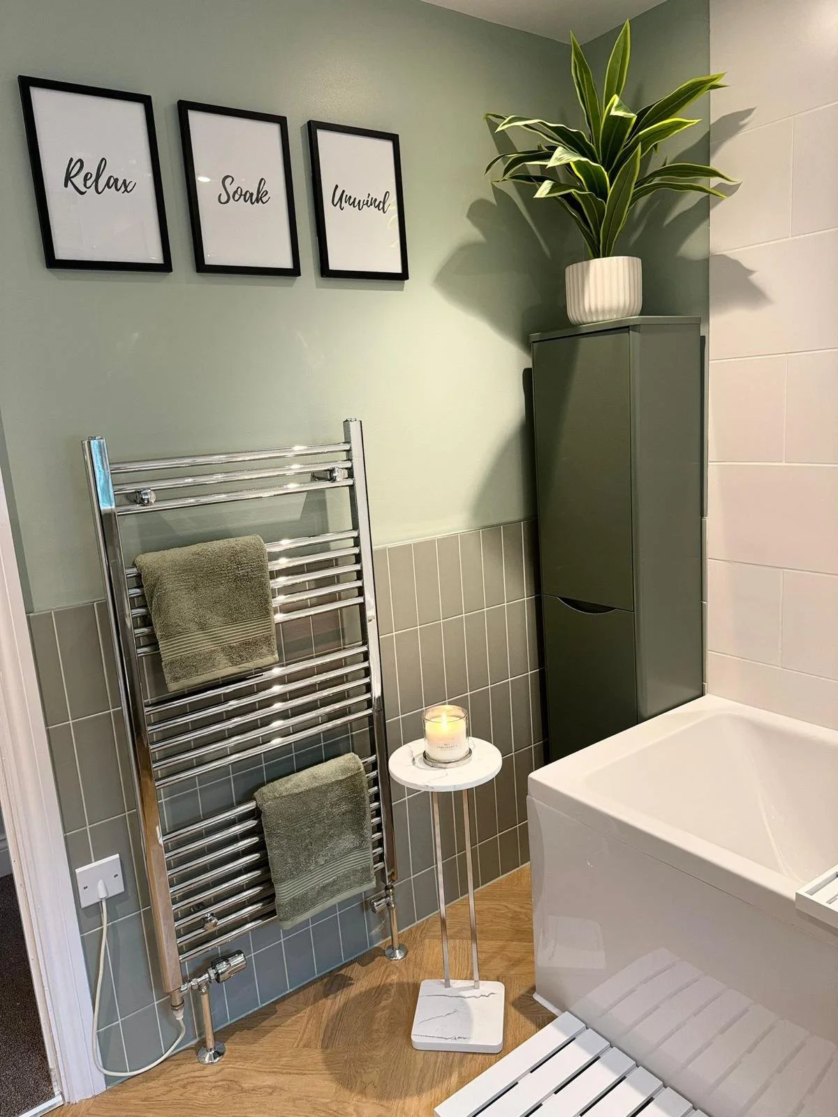 A bathroom with three framed pictures on the wall reading "Relax," "Soak," and "Unwind." There is a tall green cabinet with a potted plant on top, a white bathtub, a small white side table with a lit candle, a chrome towel radiator with two green tow