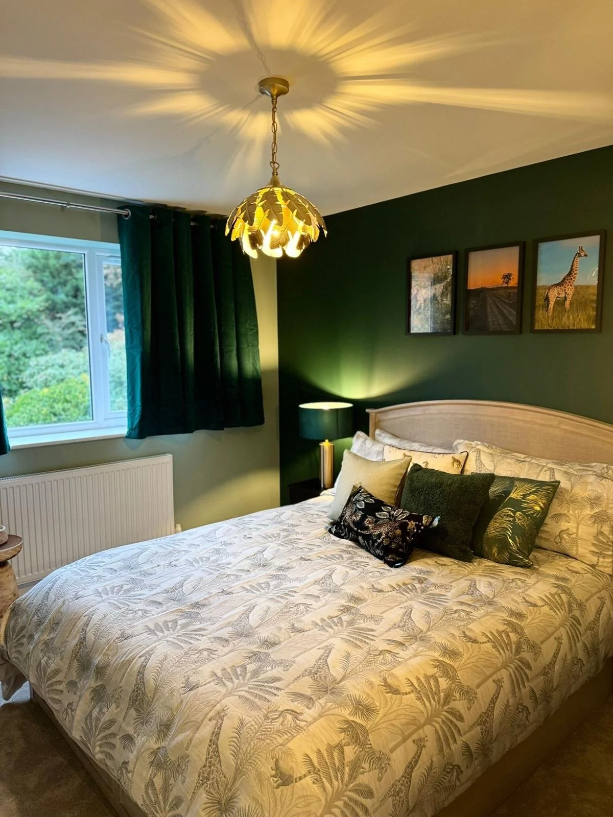 A bedroom with a bed covered in patterned bedding, multiple decorative pillows, a bedside lamp, dark green curtains, a window showing greenery outside, and framed animal pictures on a dark green wall. A ceiling light fixture casts a decorative shadow
