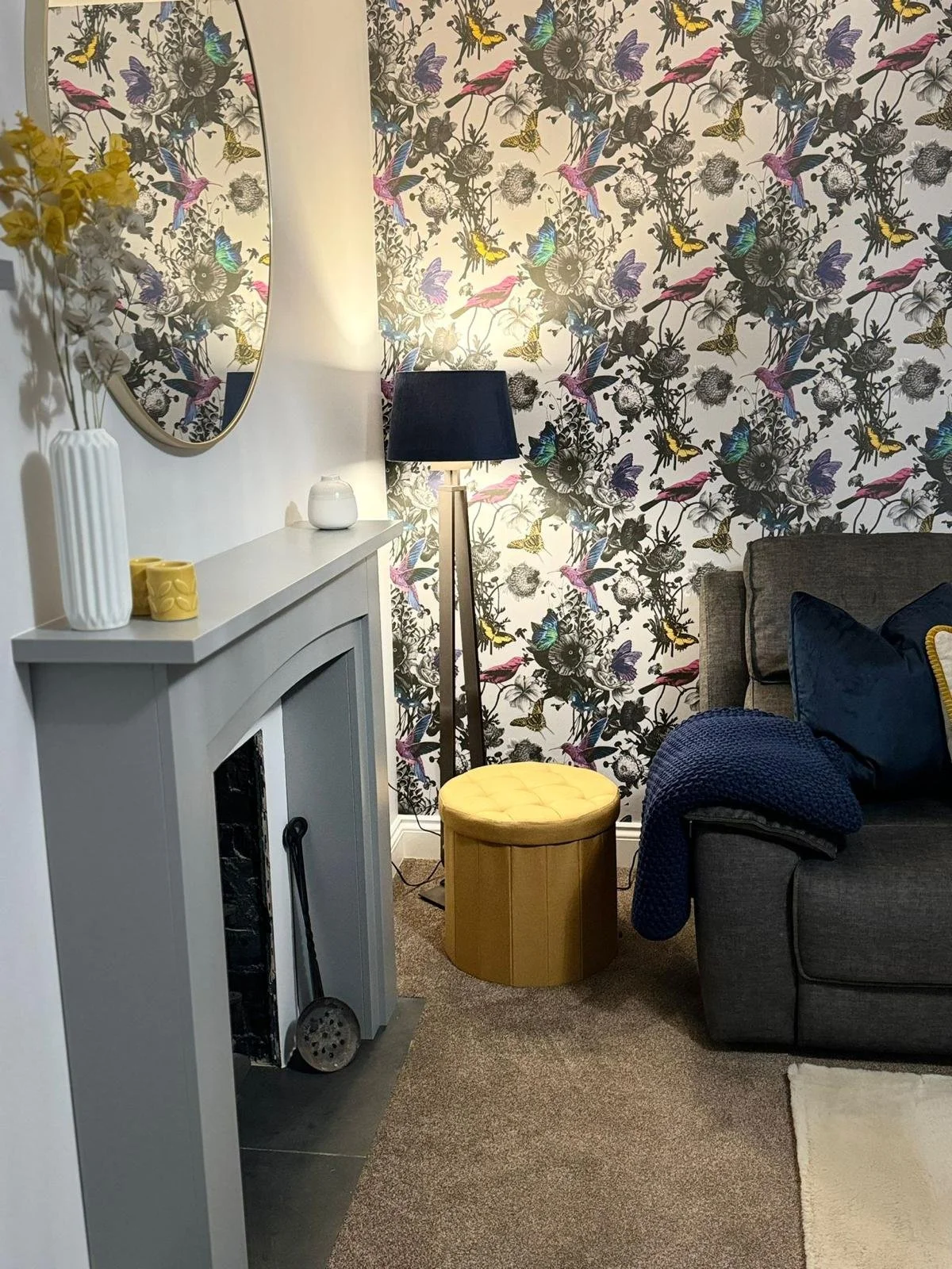 Living room corner with a gray sofa, a yellow ottoman, a black floor lamp, a floral wallpaper, a gray fireplace mantle with a white vase, yellow cups, and a mirror, beige carpet, and a navy blue blanket on the sofa.