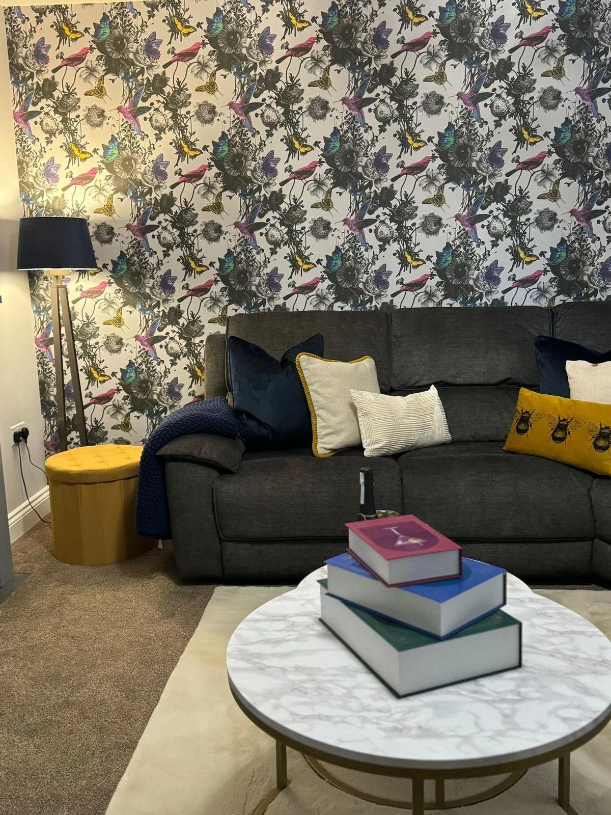 A living room featuring a black sofa with decorative pillows, a round white marble coffee table with stacked books, a yellow ottoman, a tall floor lamp with a black lampshade, and a floral patterned wallpaper with birds and flowers.