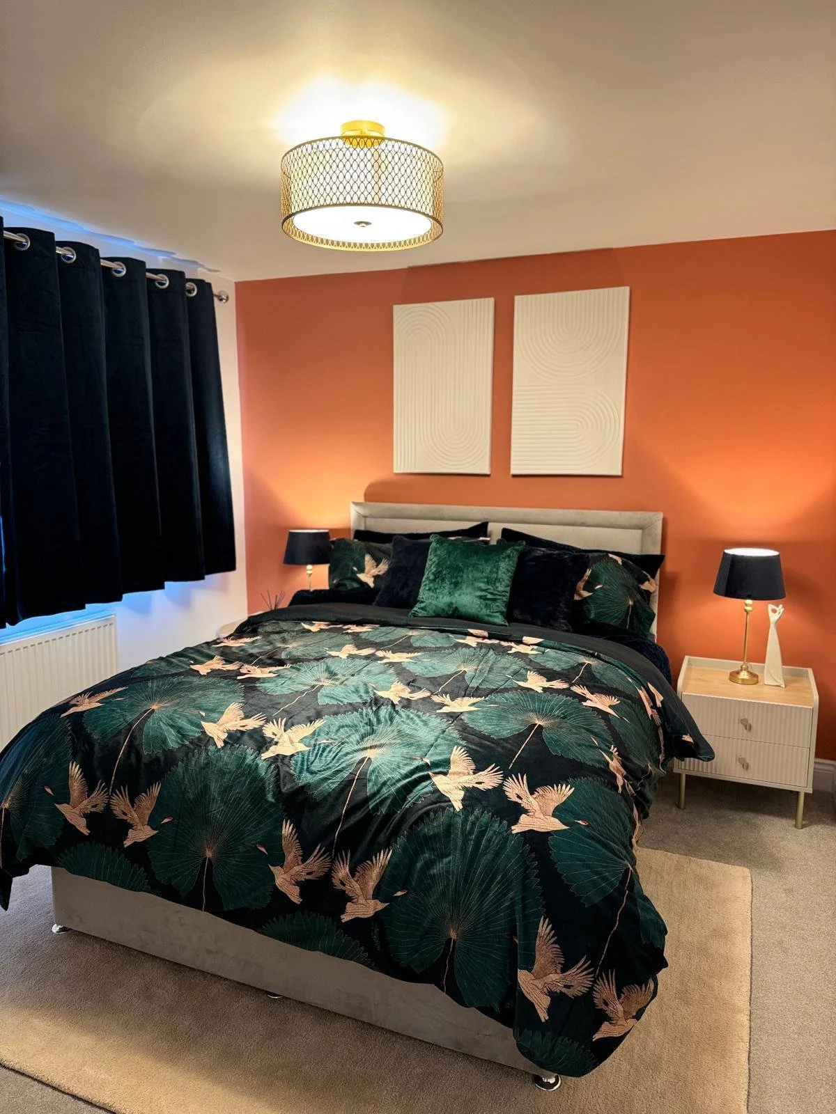 A bedroom with a peach-colored accent wall behind a bed, which has a beige upholstered headboard. The bed is made with a dark green and black bird-themed comforter and matching pillows. On each side of the bed, there are small white nightstands with 