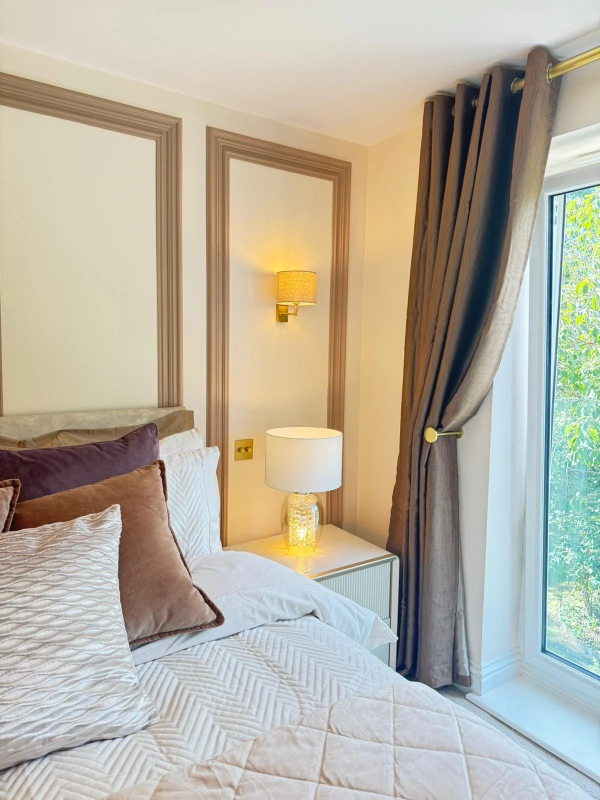 A cozy bedroom corner with a bed, multiple pillows, a white bedside table with a lamp, beige wall with decorative molding, a wall-mounted lamp, and a window with brown curtains and a view of green foliage outside.