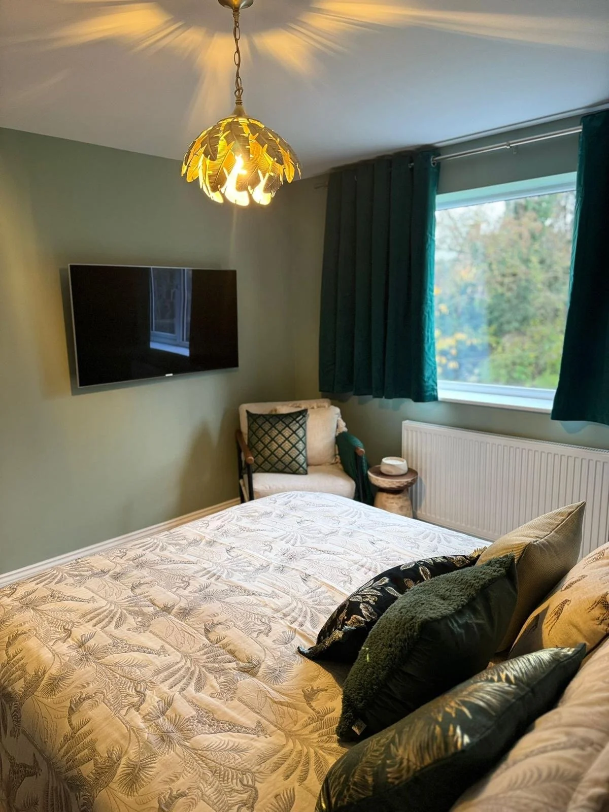 A cozy bedroom with a bed, decorative pillows, a wall-mounted flat-screen TV, a window with teal curtains, a chair with a cushion, a small side table, and a ceiling light with a leafy design.