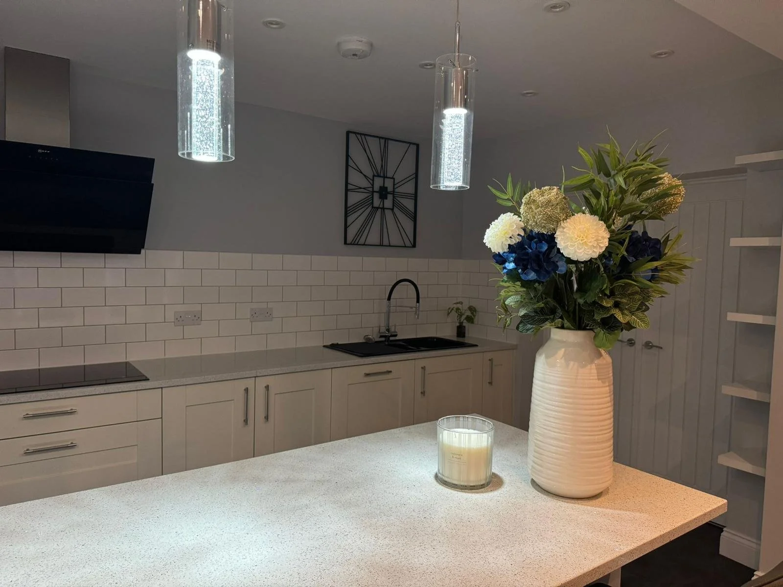 A modern kitchen with white cabinets, a black stovetop, a black sink, and white subway tile backsplash. A large white vase with a mixed floral arrangement and a candle on the counter are in the foreground, with hanging pendant lights overhead.