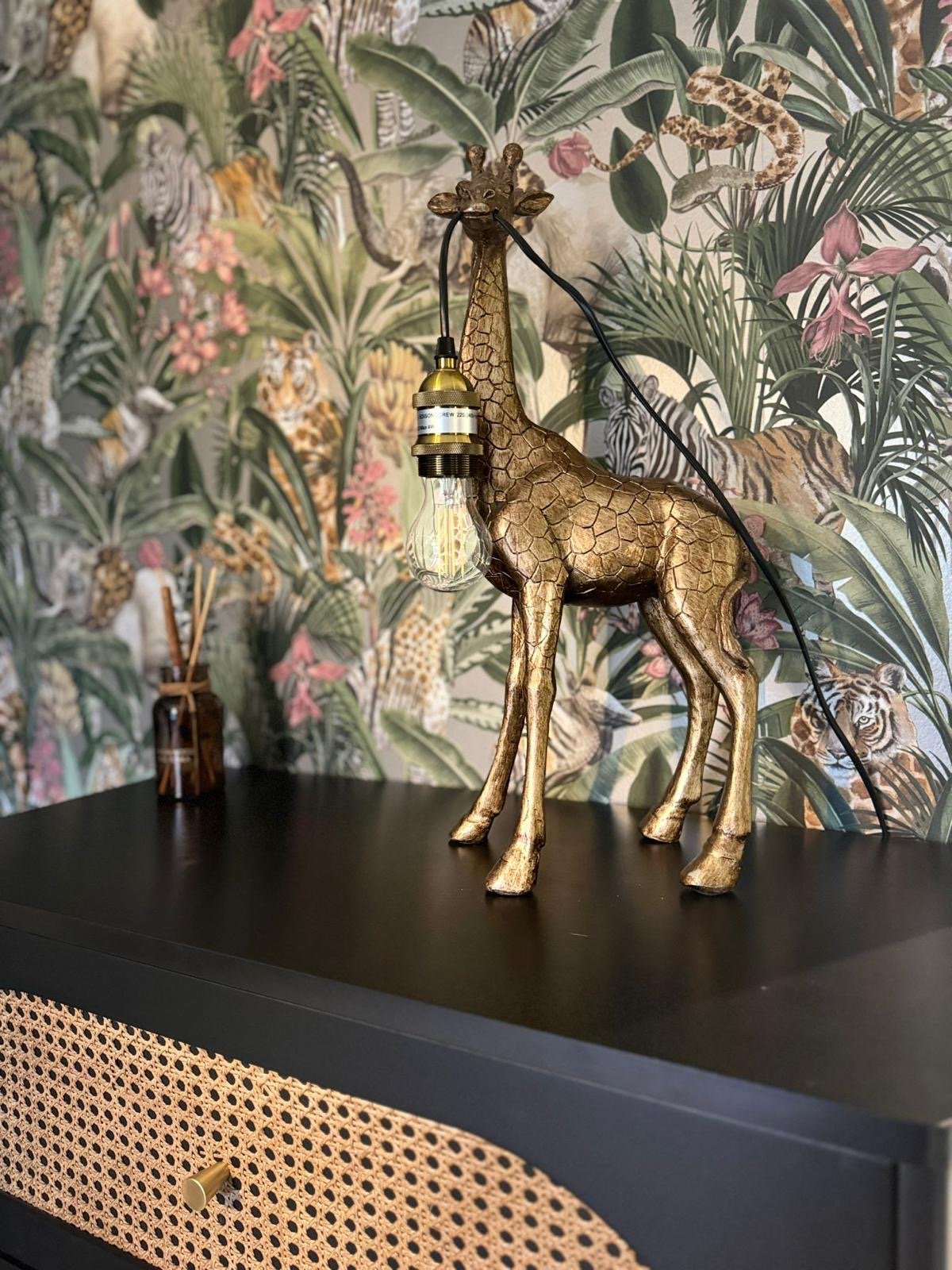 A decorative gold-painted giraffe lamp with a light bulb hanging from its neck on a black table, with a jungle-themed wallpaper featuring animals and tropical foliage in the background.