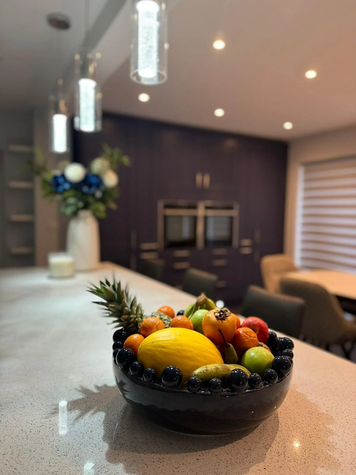 Bowls of mixed fruits, including pineapple, melon, and berries, on kitchen countertop with modern decor and dining area in background.