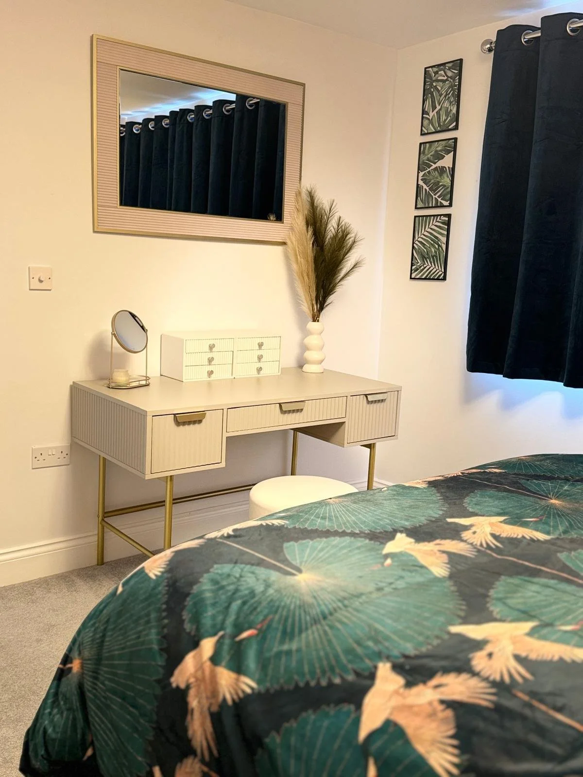 A bedroom corner with a beige desk, a small round mirror, a white stool, a tall white vase with dried pampas grass, and framed botanical prints on the wall. A mirror reflects dark curtains, and part of a bed with a green and gold patterned cover is v