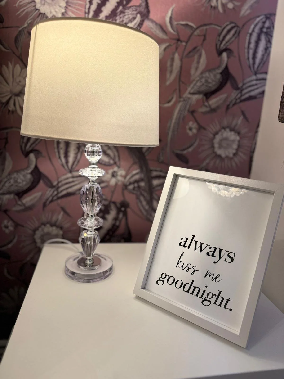 A white table with a crystal table lamp and a framed sign that reads "always kiss me goodnight" against a pink floral wallpaper background.