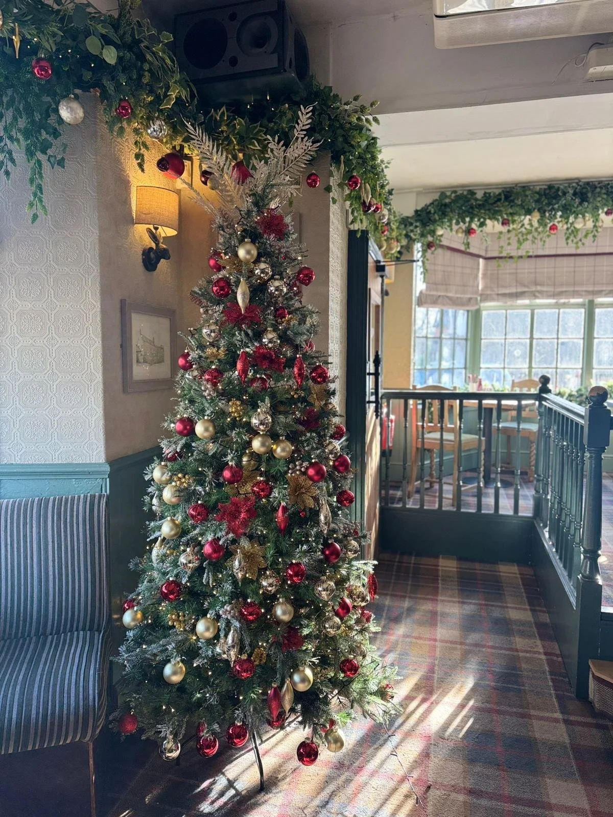 Decorated Christmas tree with red and gold ornaments in a cozy restaurant or cafe with natural light, garland, and holiday decorations.