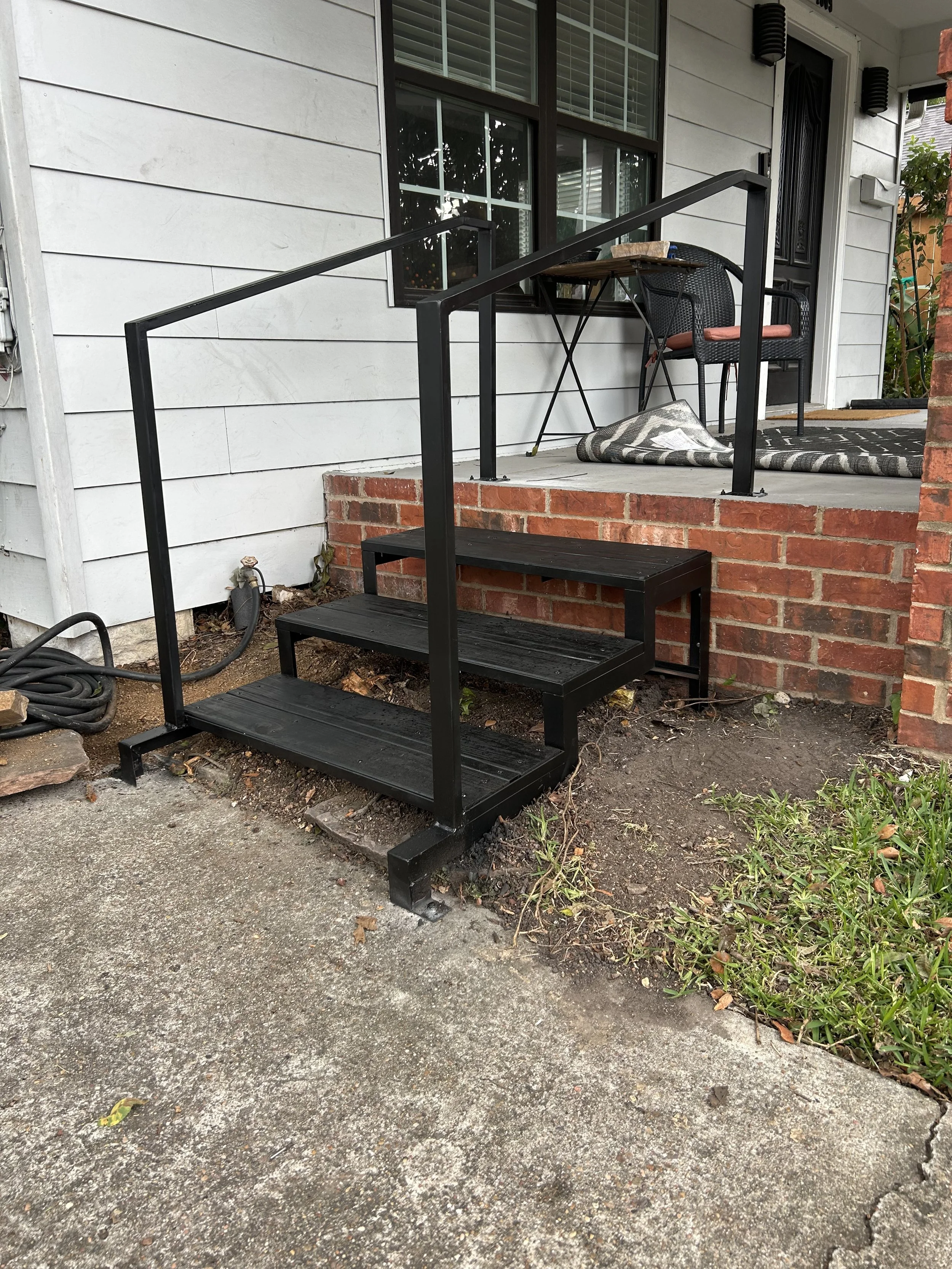 Black metal outdoor staircase with two steps leading up to a porch with white siding, brick foundation, and patio furniture.