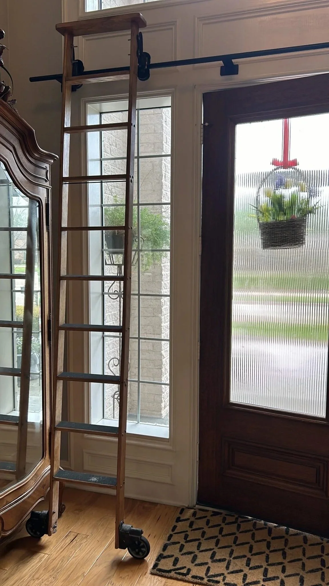A wooden ladder with wheels positioned inside a house near a front door with a hanging basket of flowers outside the glass door.