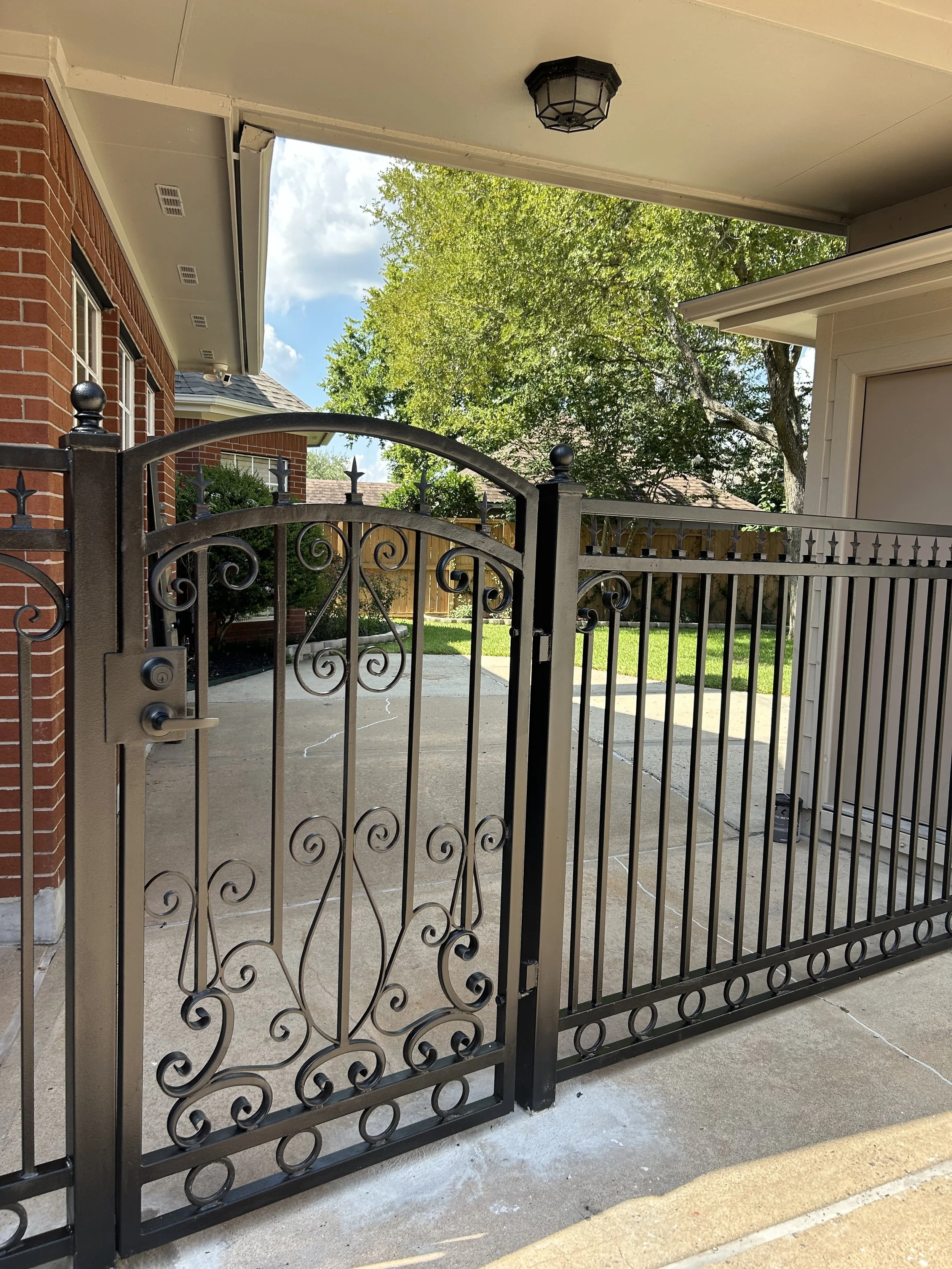 Black wrought iron gate with decorative swirls in front of a driveway, with a brick house on the left and a white garage on the right, green trees in the background, under a partly cloudy sky.