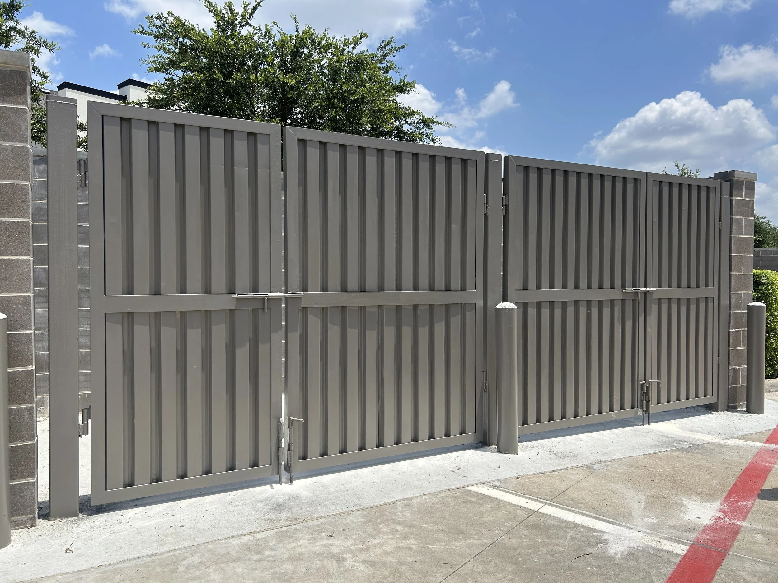 Gray metal double gates with vertical slats, supported by brick and metal posts, under a blue sky with white clouds.