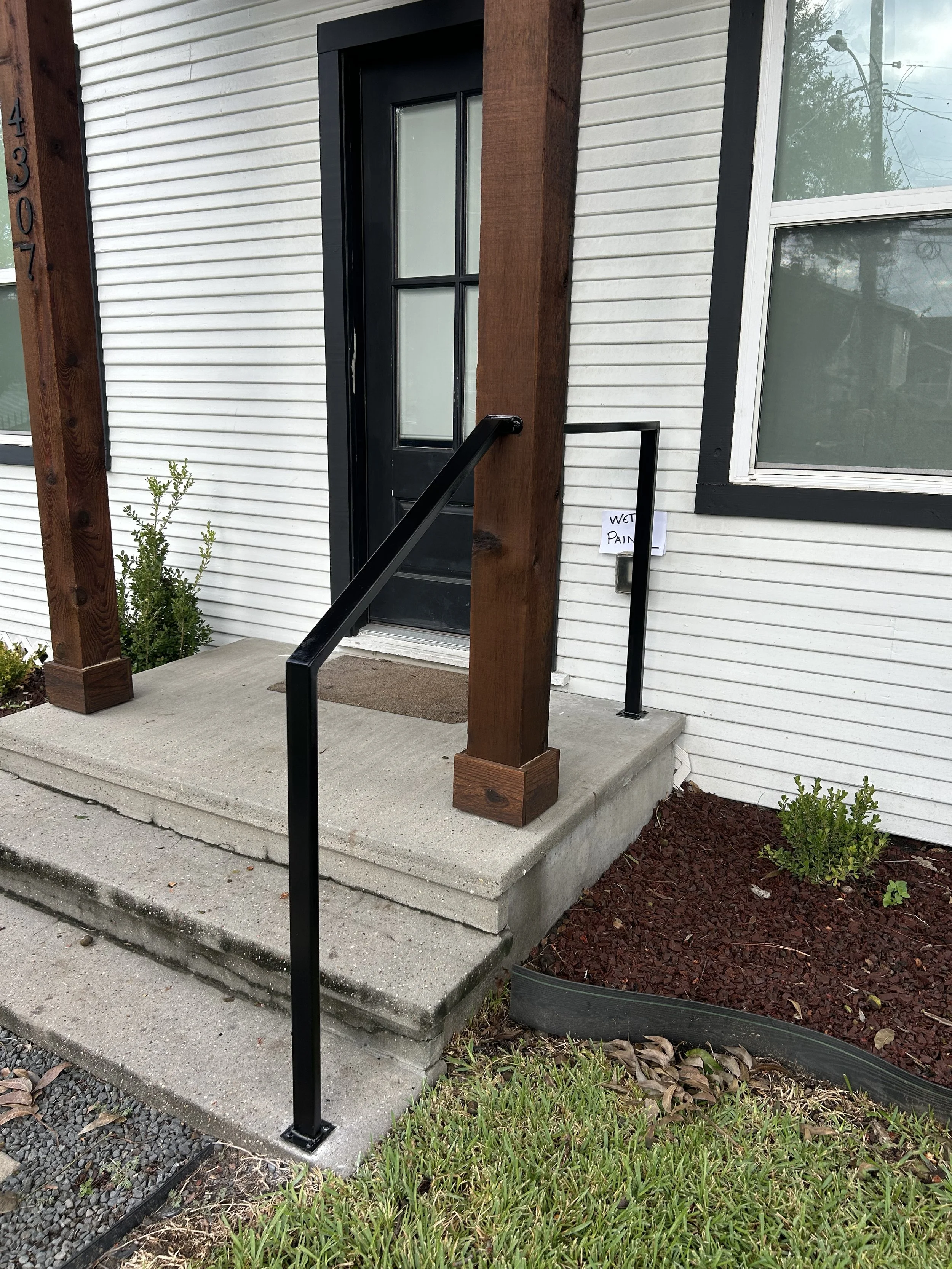 The image shows the front porch of a house with a black door, a concrete step, and a black metal handrail. The house has white siding with black trim around the window, and there are small plants and mulch beds on the sides of the porch. A small hand