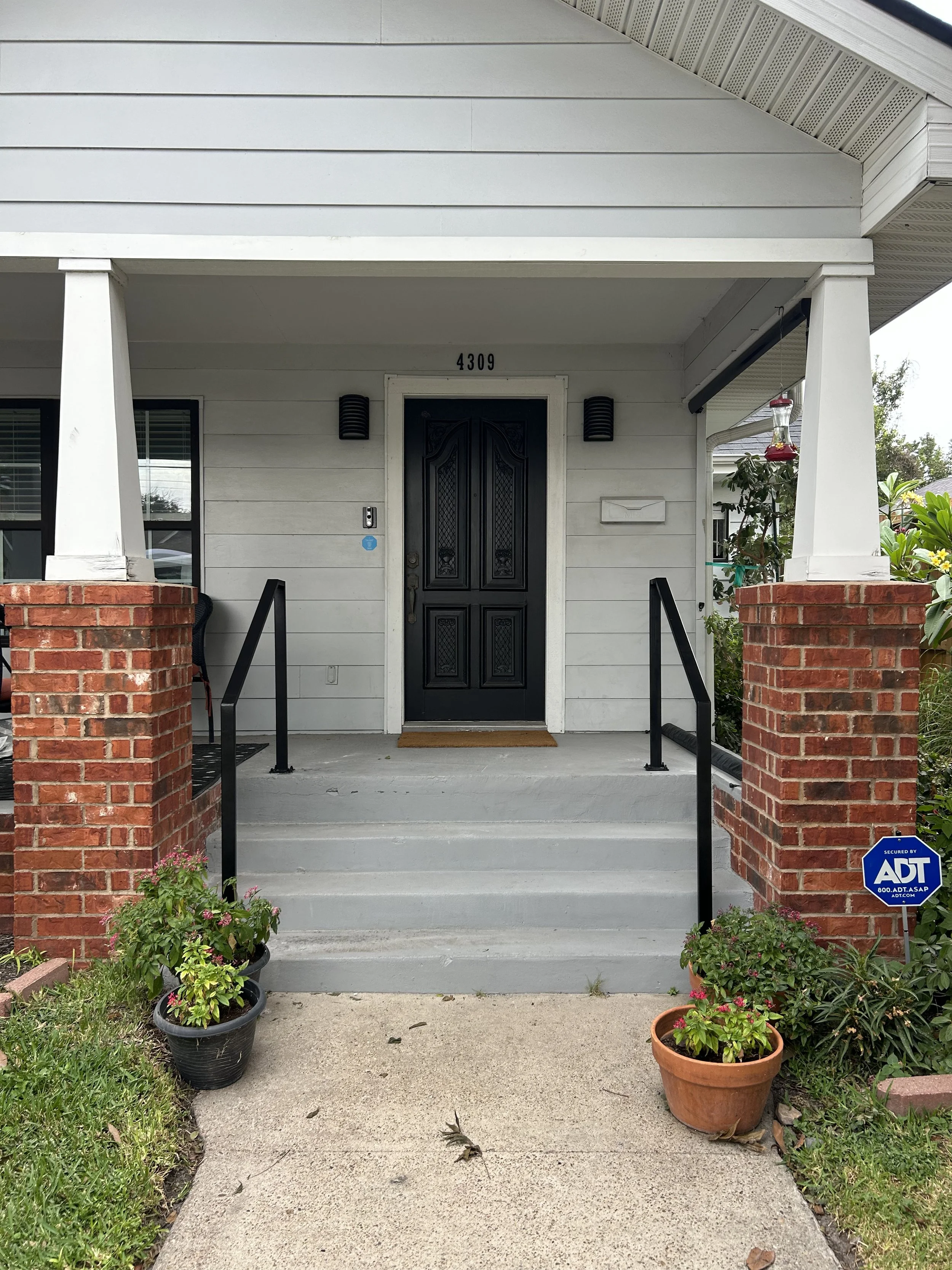 Front porch of a house with white walls, black door, brick pillars, potted plants, and security sign.