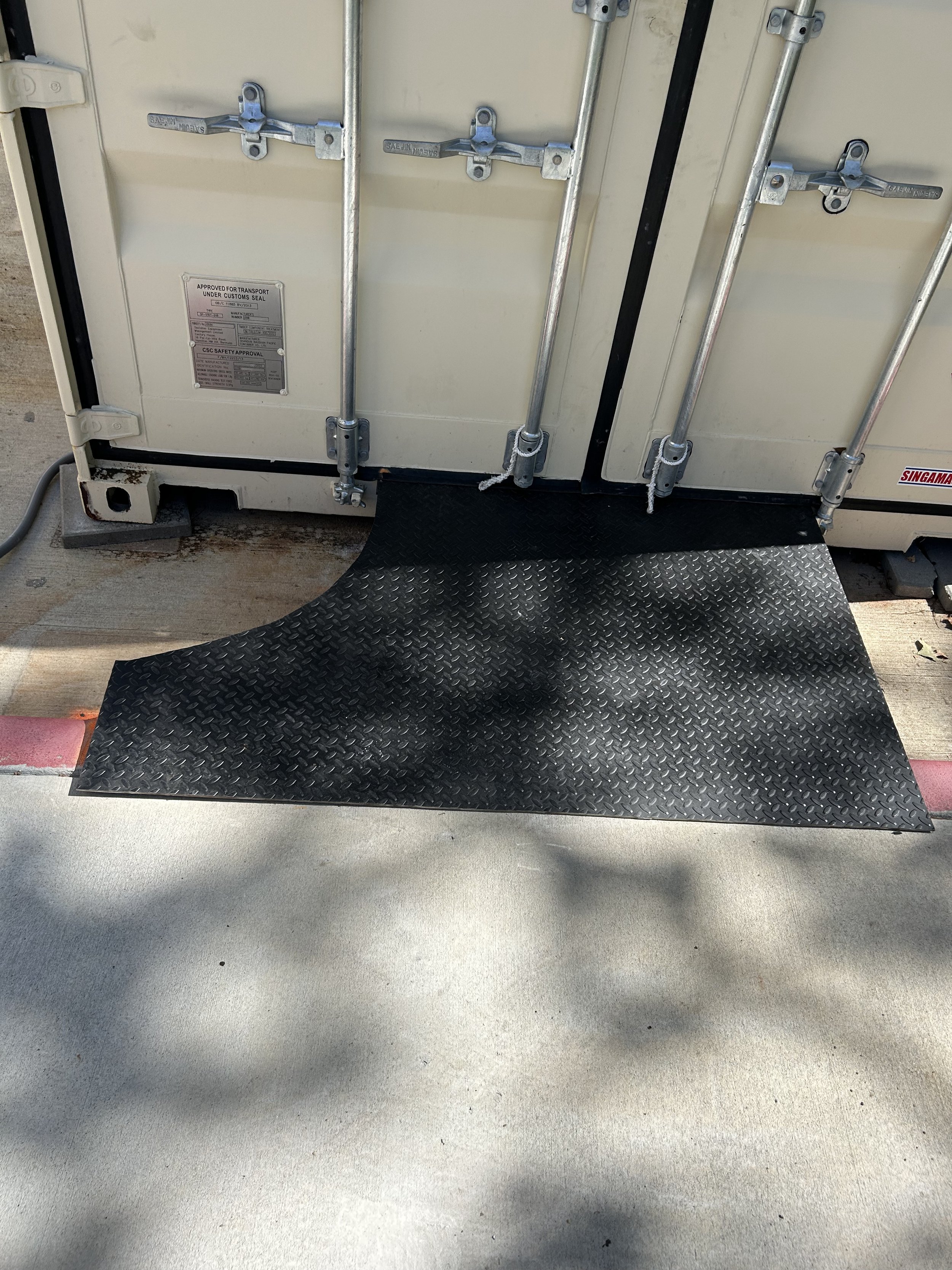 A black rubber ramp extending from a white industrial container, with parking lot pavement and shadows visible.