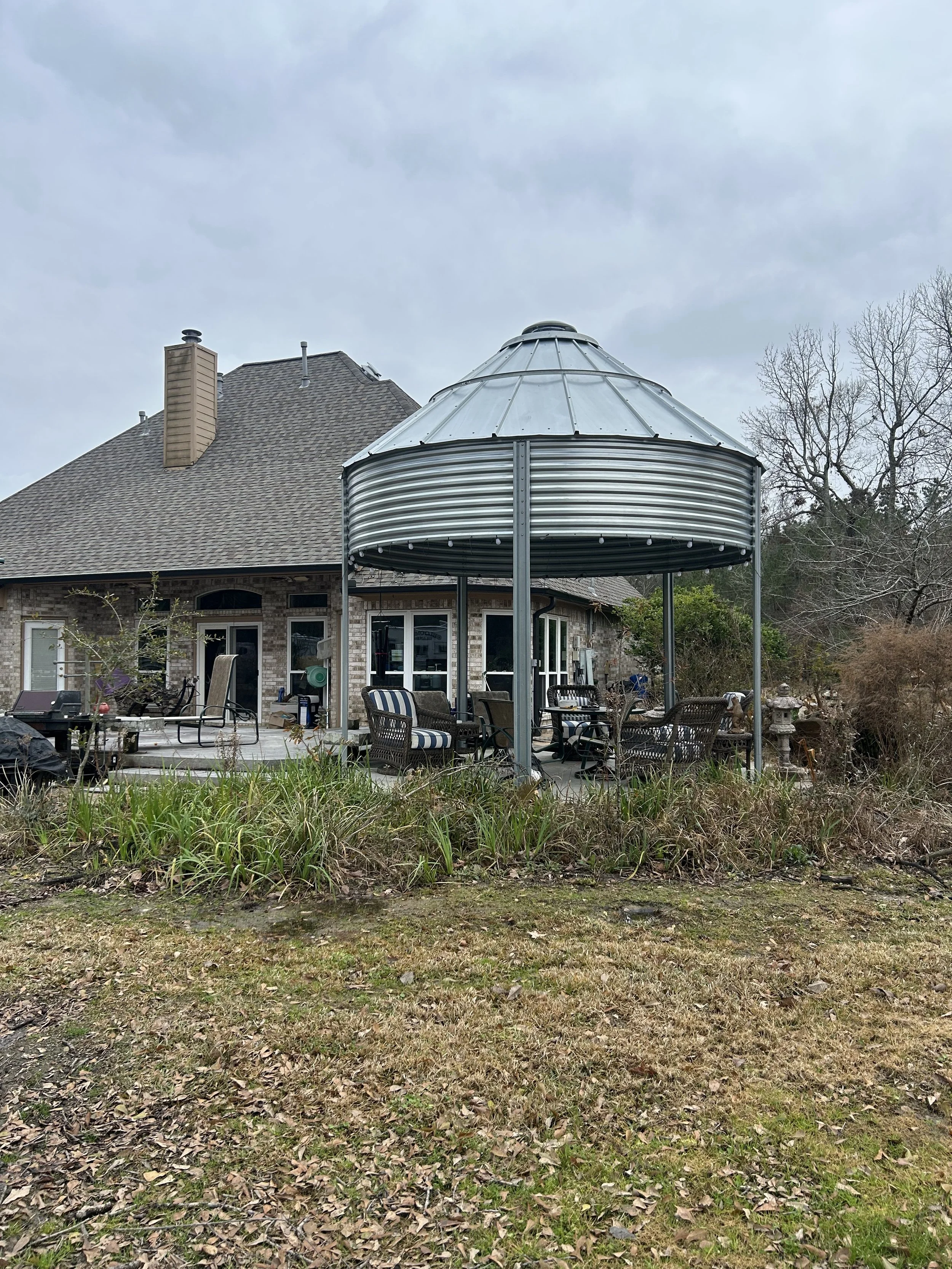Backyard with patio furniture under a metal gazebo, house with brick exterior, leafless trees, and cloudy sky.