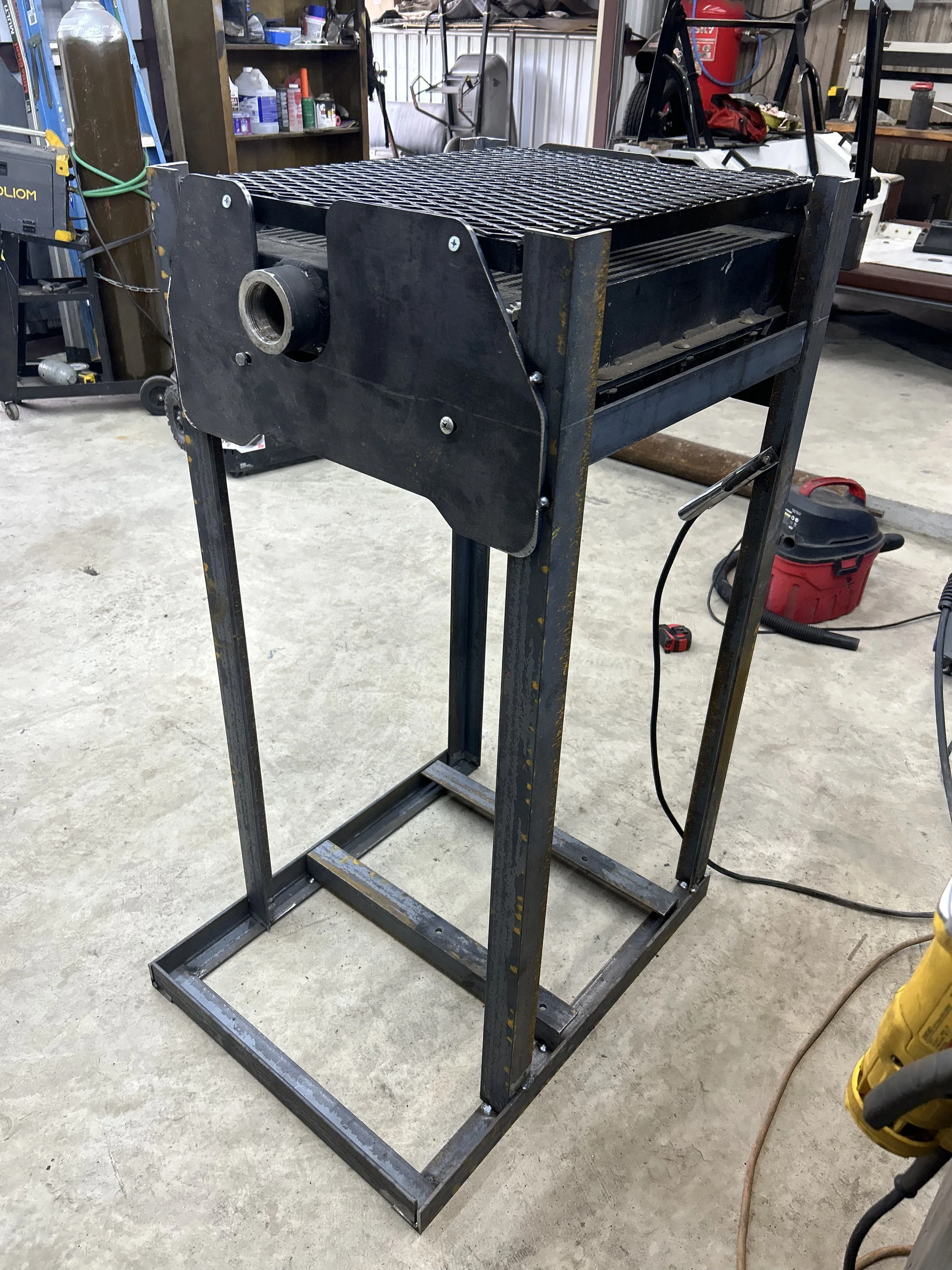 Metal work table with a grated top in a workshop setting, equipped with a frame and some tools and shelves in the background.