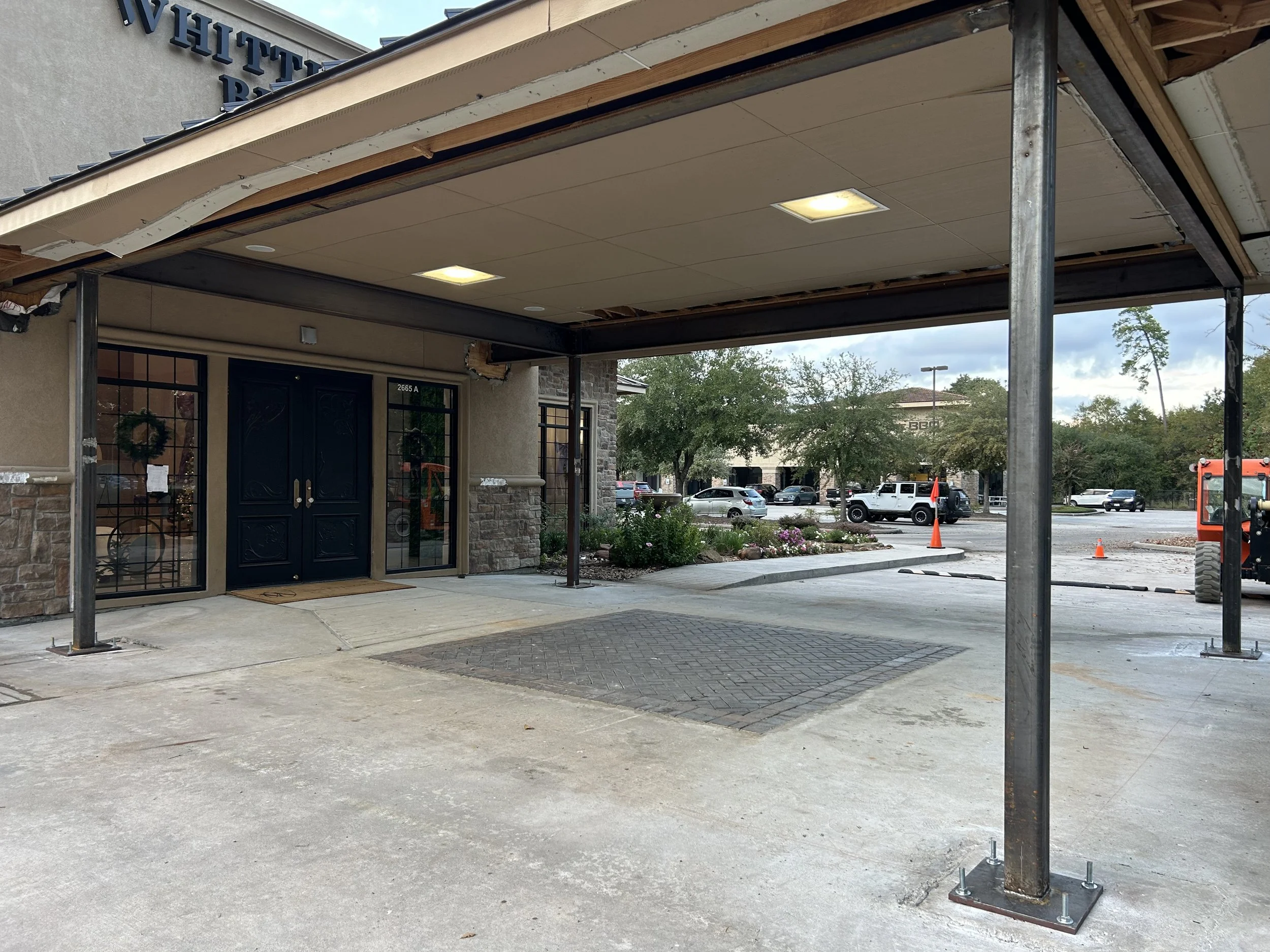 Building under construction with a covered entrance, black front door, large windows, and parking lot with construction cones and equipment in the background.