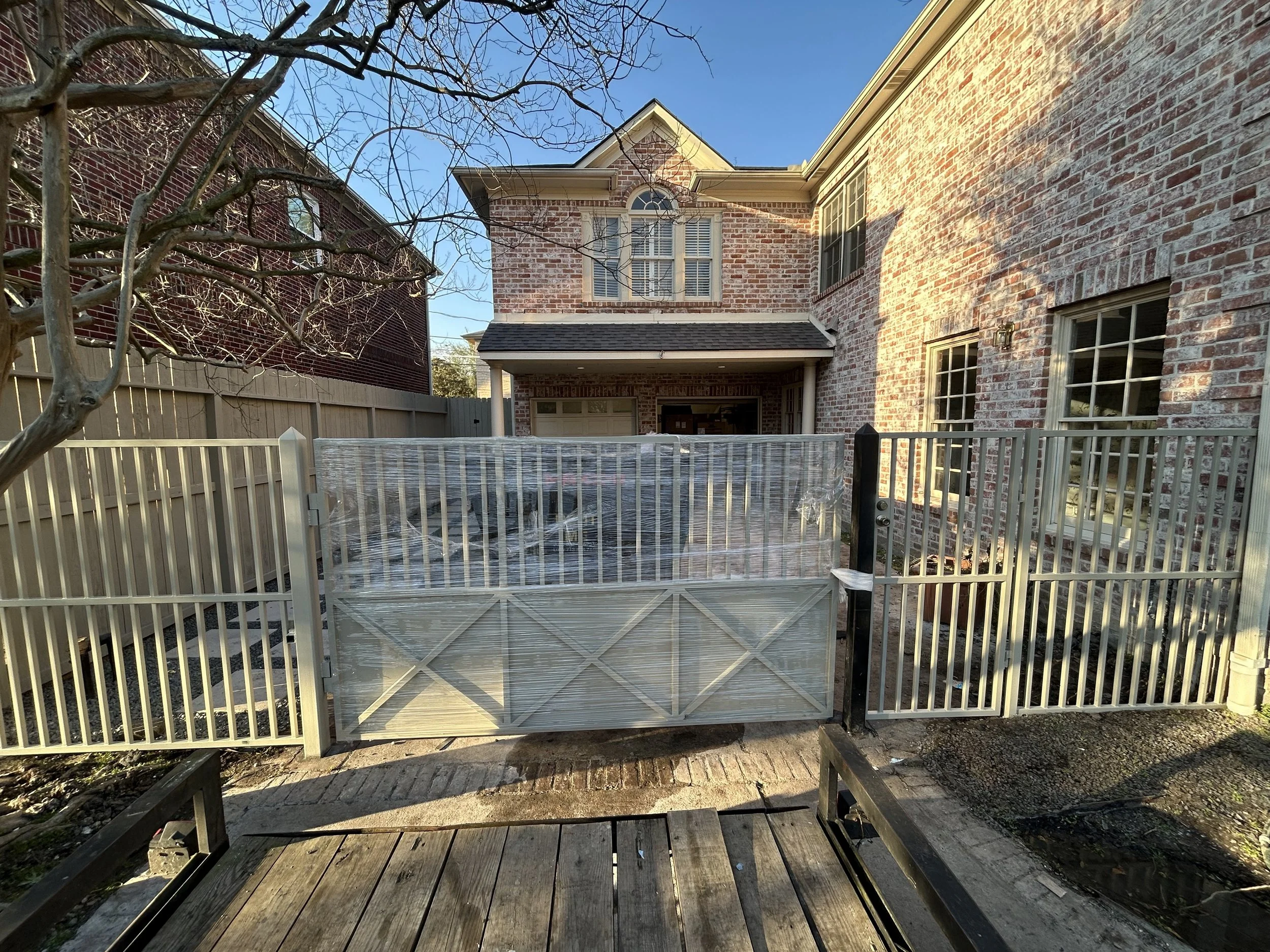 The backyard of a brick house with a white fence and gate, a tree with bare branches, and a porch area visible in the background. The fence has a gate wrapped in plastic.