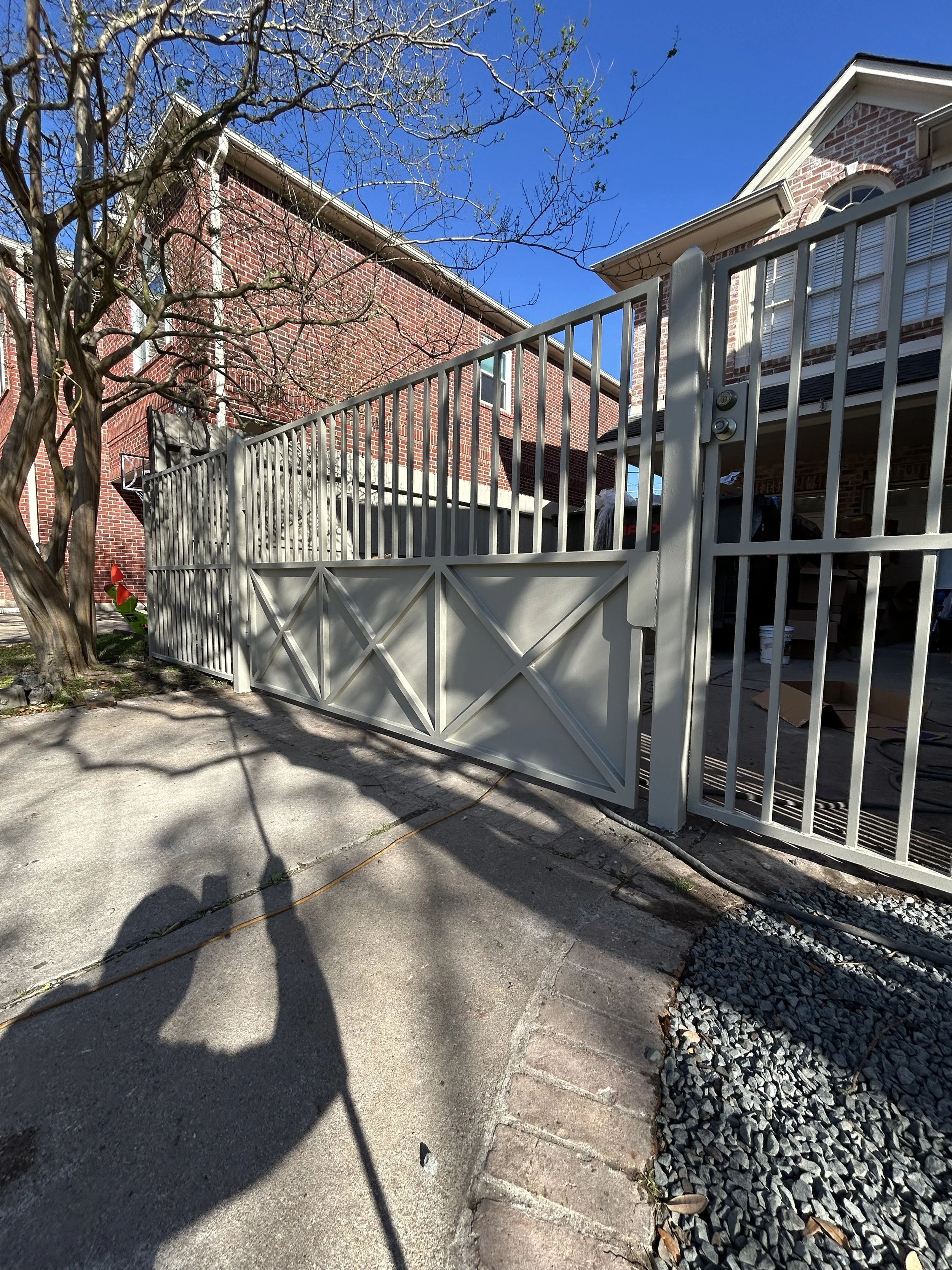 White metal fence and gate in front of a brick house with a garage. Shadows cast on the driveway, with leafless trees and a clear blue sky.